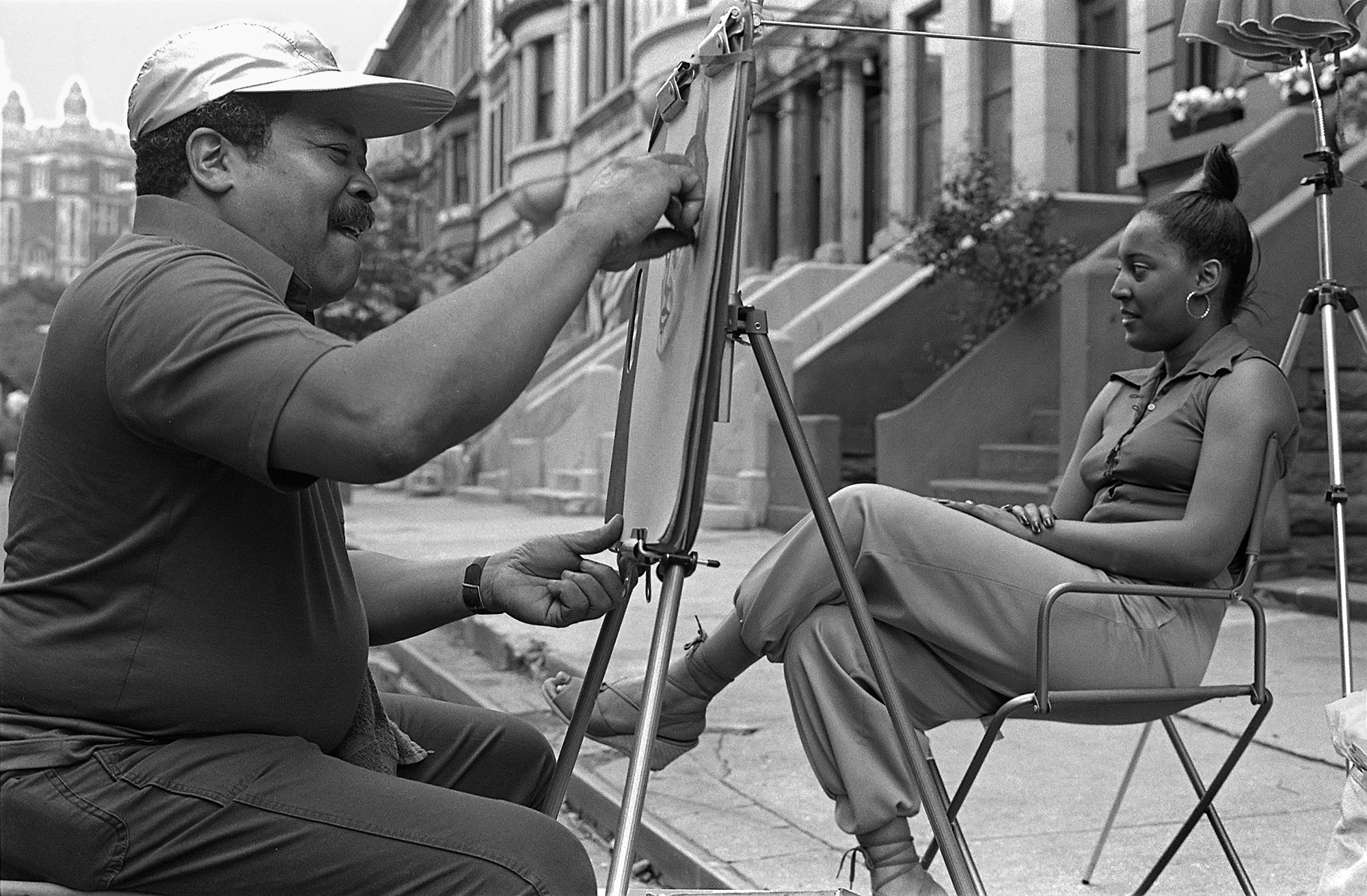 a black and white photo of a street artist drawing a woman sitting before him in a chair