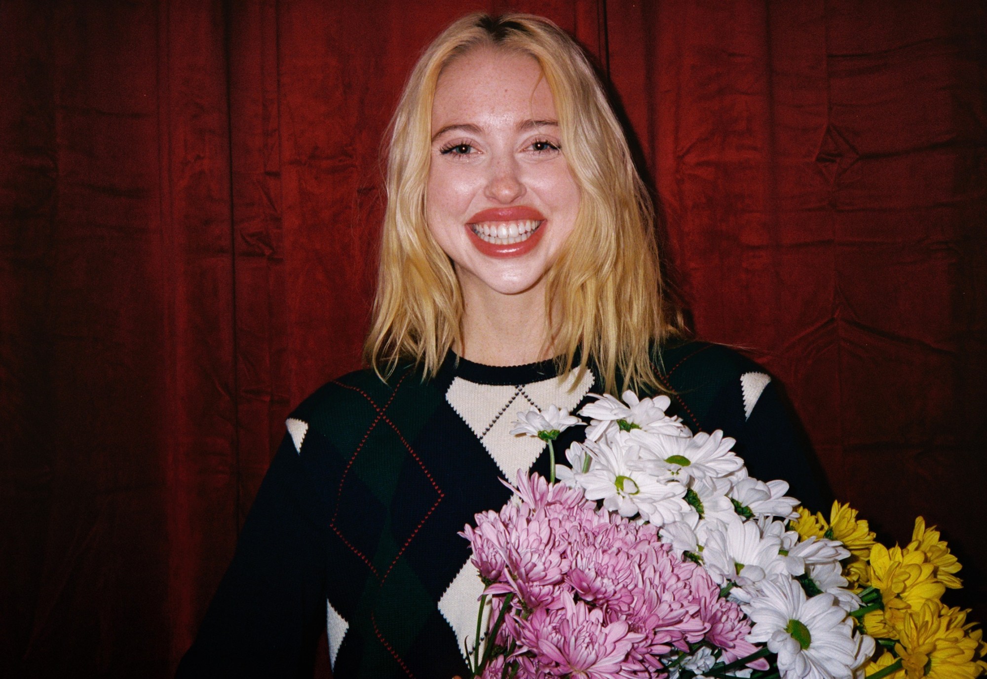 chloe cherry smiling wide wearing an argyle sweater and holding a bouquet of flowers