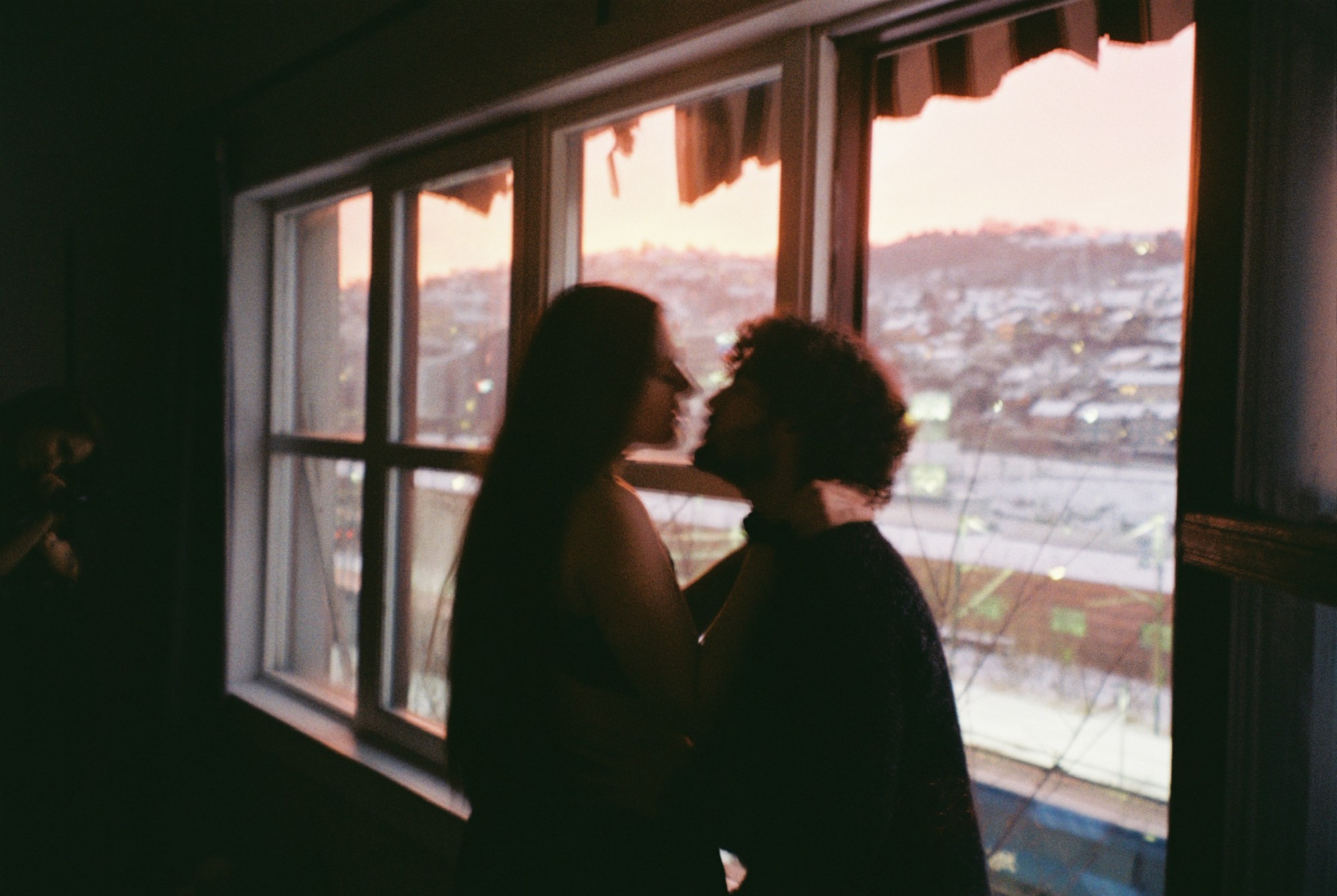 A couple kissing in a dimly lit room against a window looking out over the city.