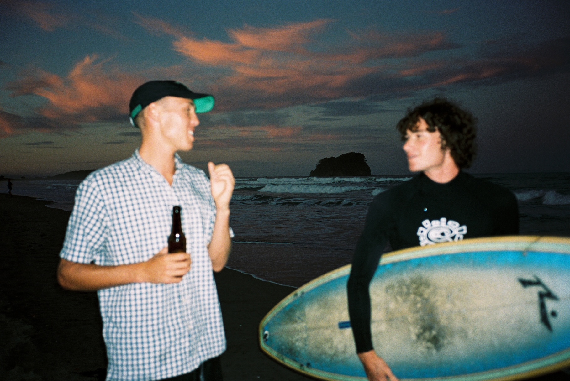 Two men on a beach in the morning. One in a shirt holds a beer, the other in a wetsuit holds a surfboard.