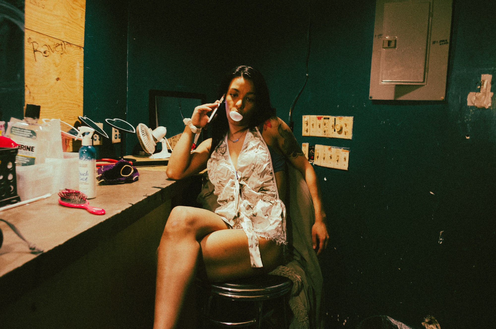 A girl blows a bubblegum bubble while sitting with her phone to her ear in a dressing room