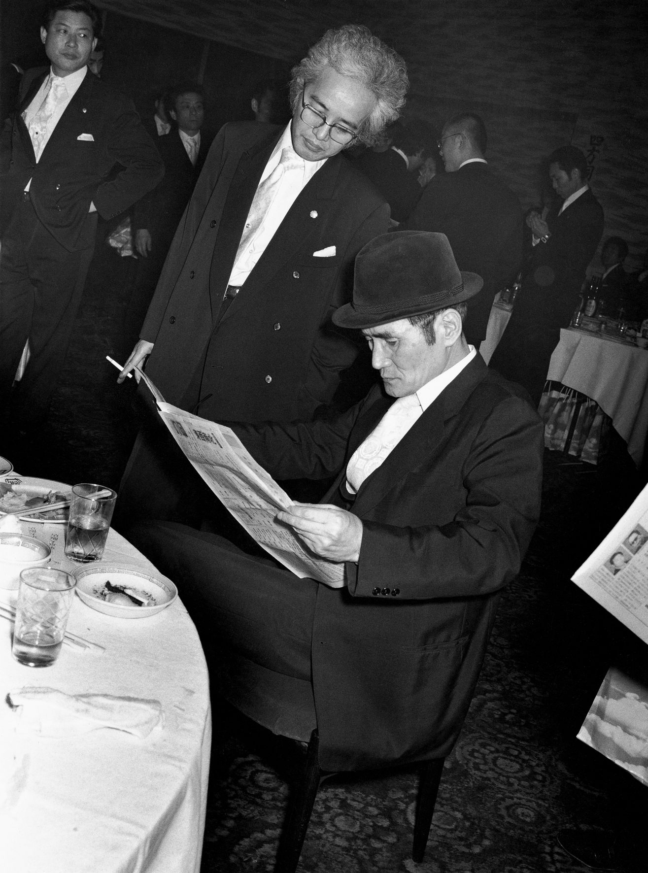 a man in a suit and hat reading the paper at a dinner table in 70s tokyo by seiji kurata