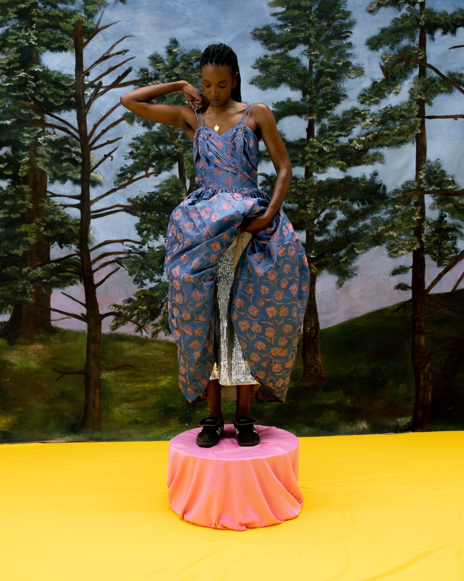 a model in a floral dress stands on a pink platform in front of a painted woodland mural