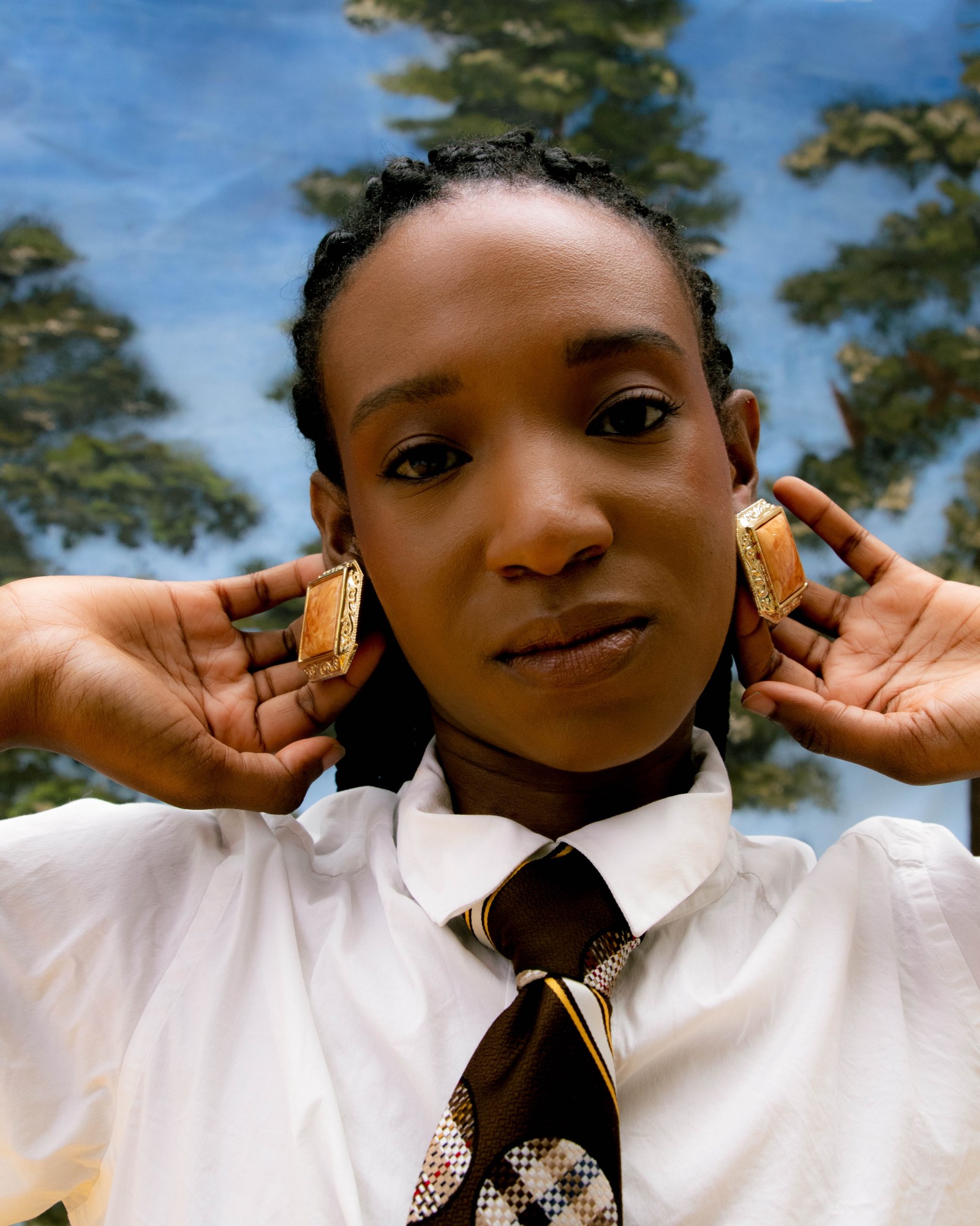 a close up of a model in a shirt and tie, showing off large earrings, in front of a painted woodland mural