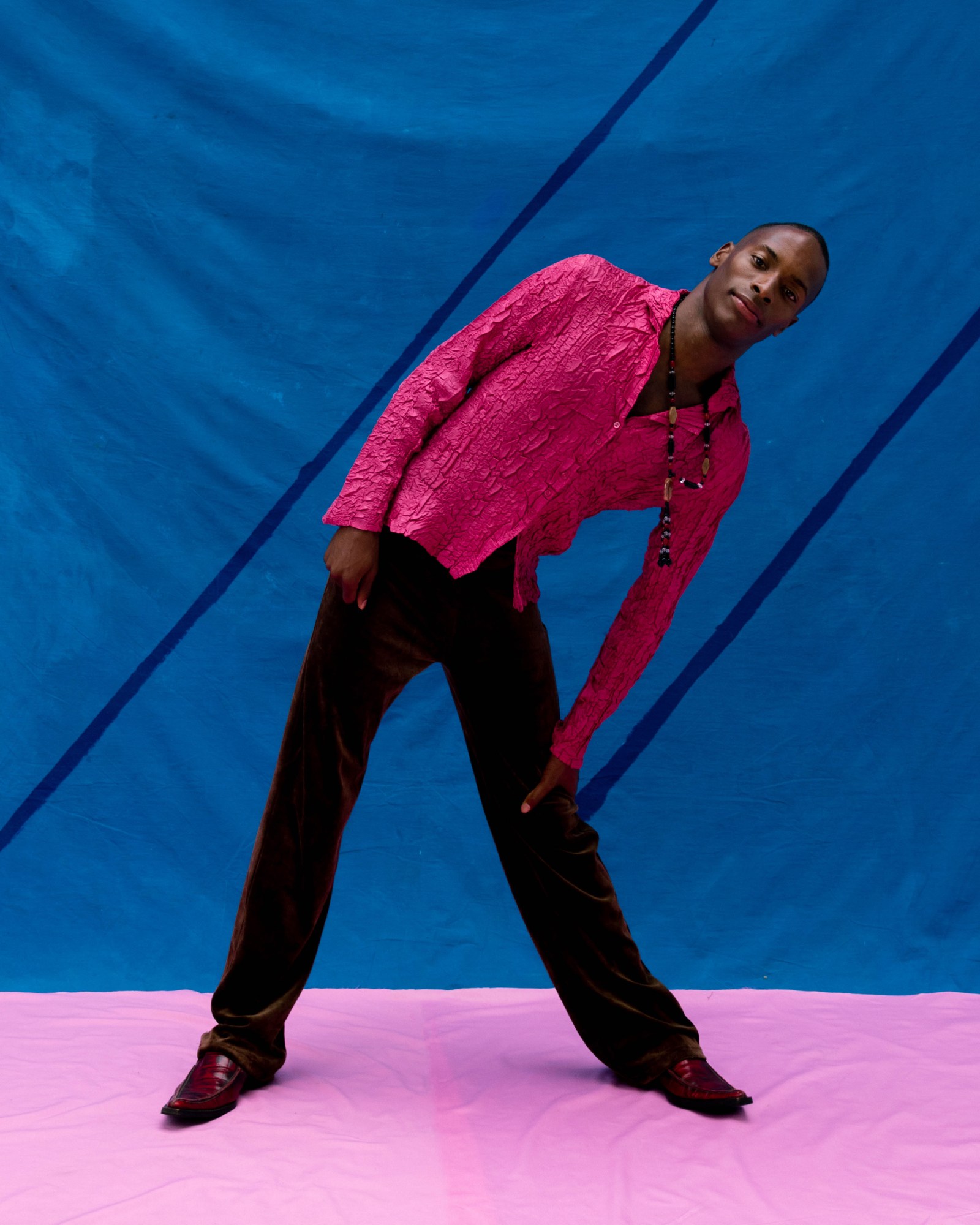 a model wearing a hot pink shirt stands leaning sideways in front of a blue painted backdrop