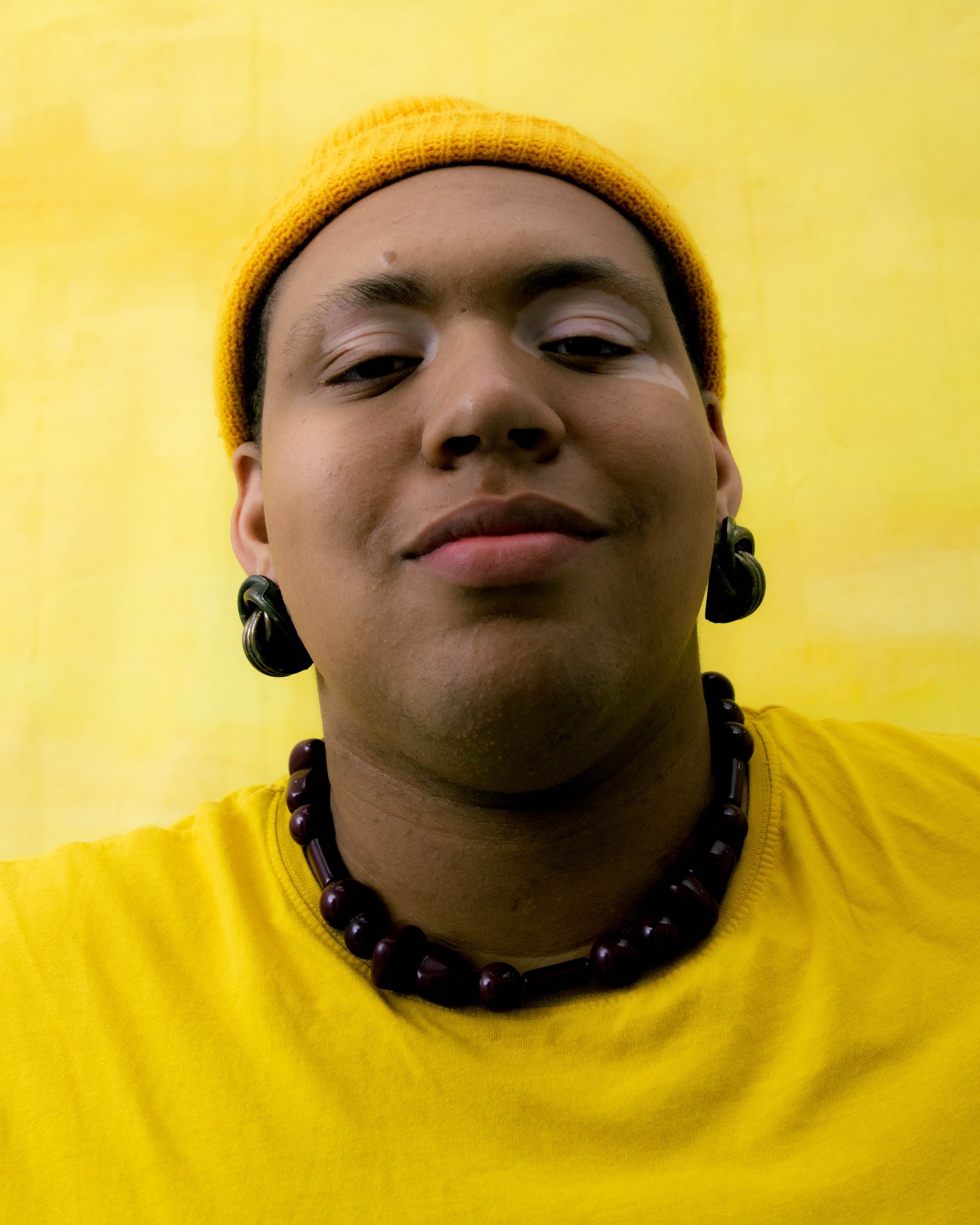 a smiling model wears a bright yellow t-shirt and beanie in front of a matching yellow backdrop