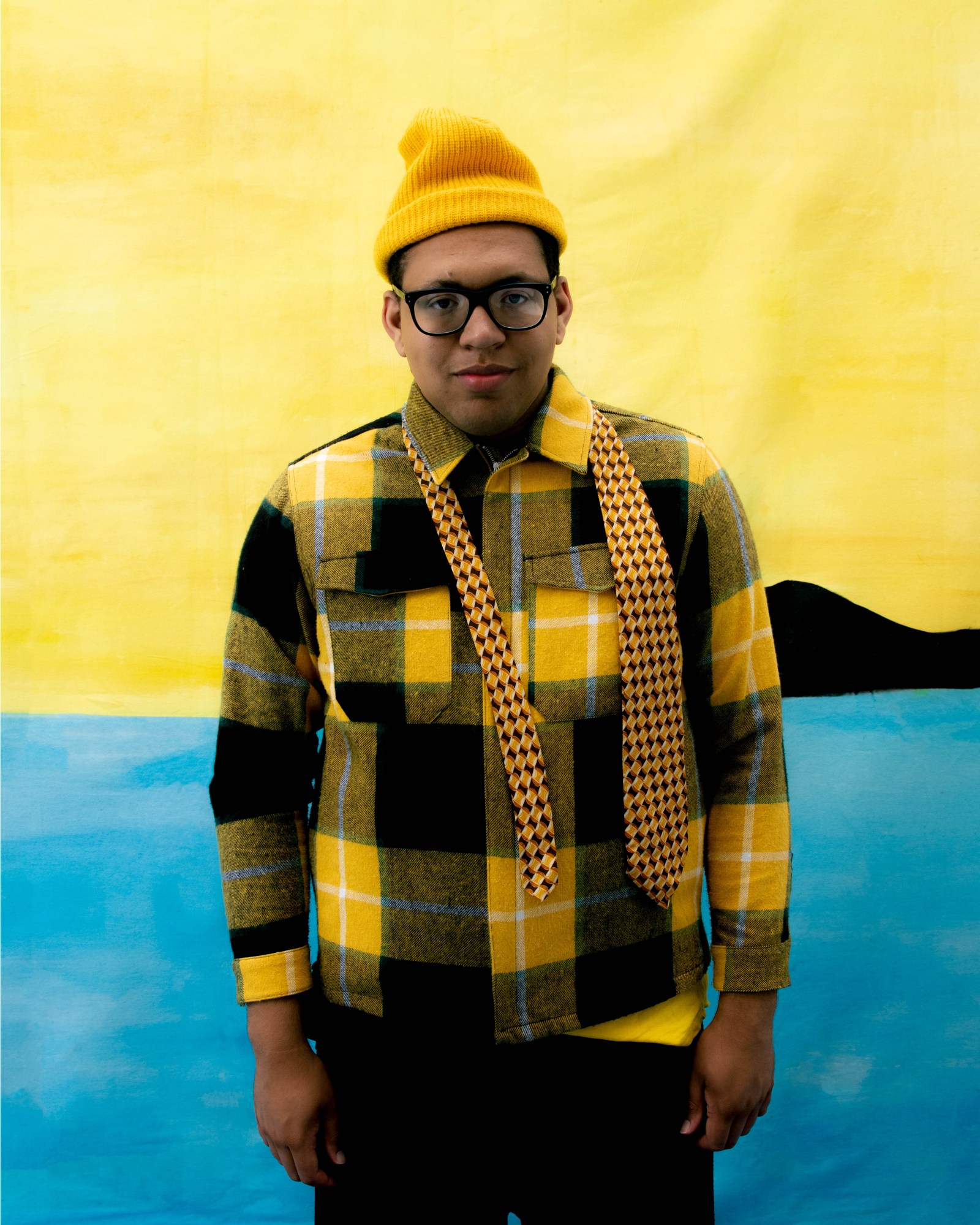 a smiling model wears a yellow t-shirt and beanie in front of a matching yellow backdrop