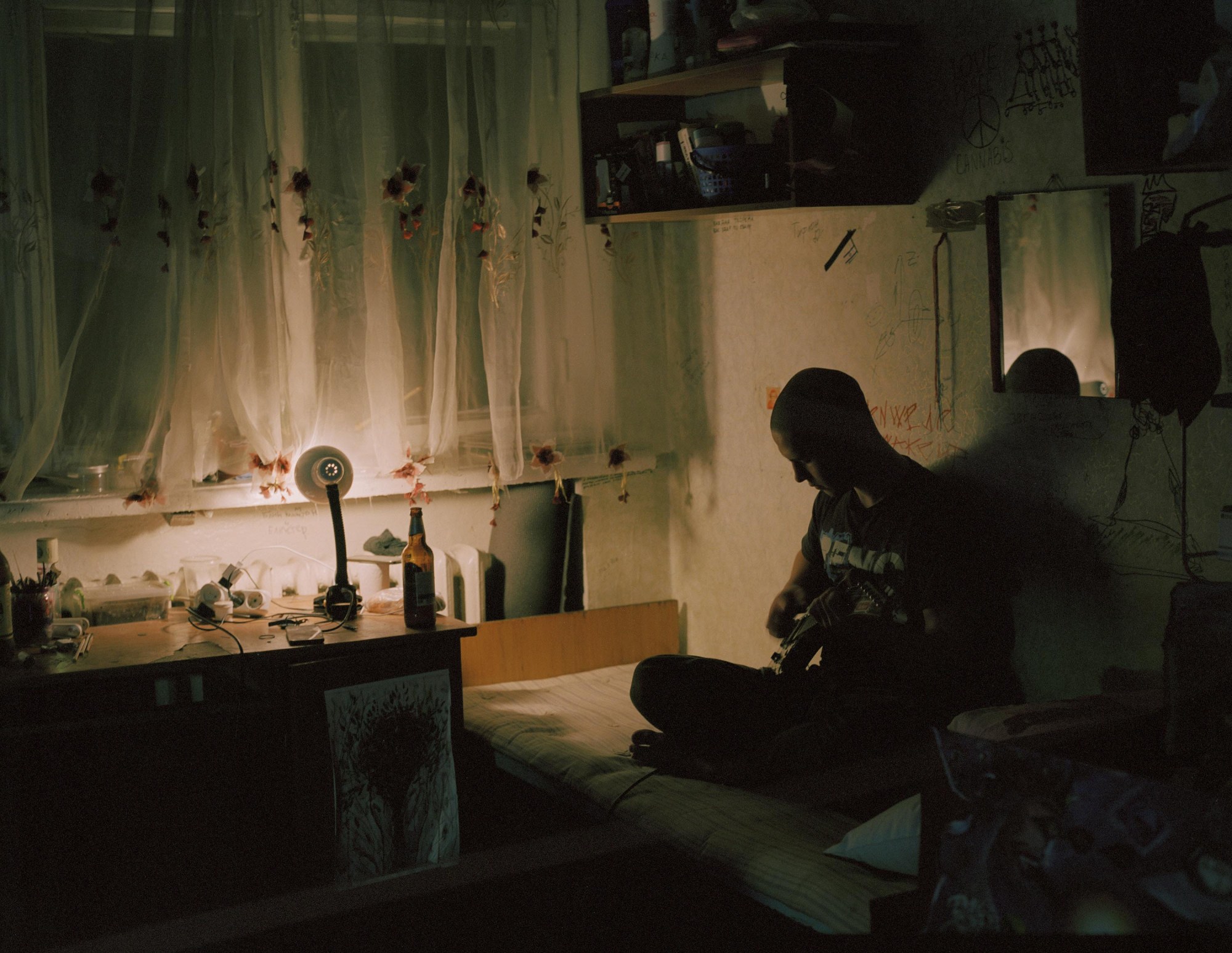 model sits silhouetted in a room lit by just a desk lamp, strumming a guitar