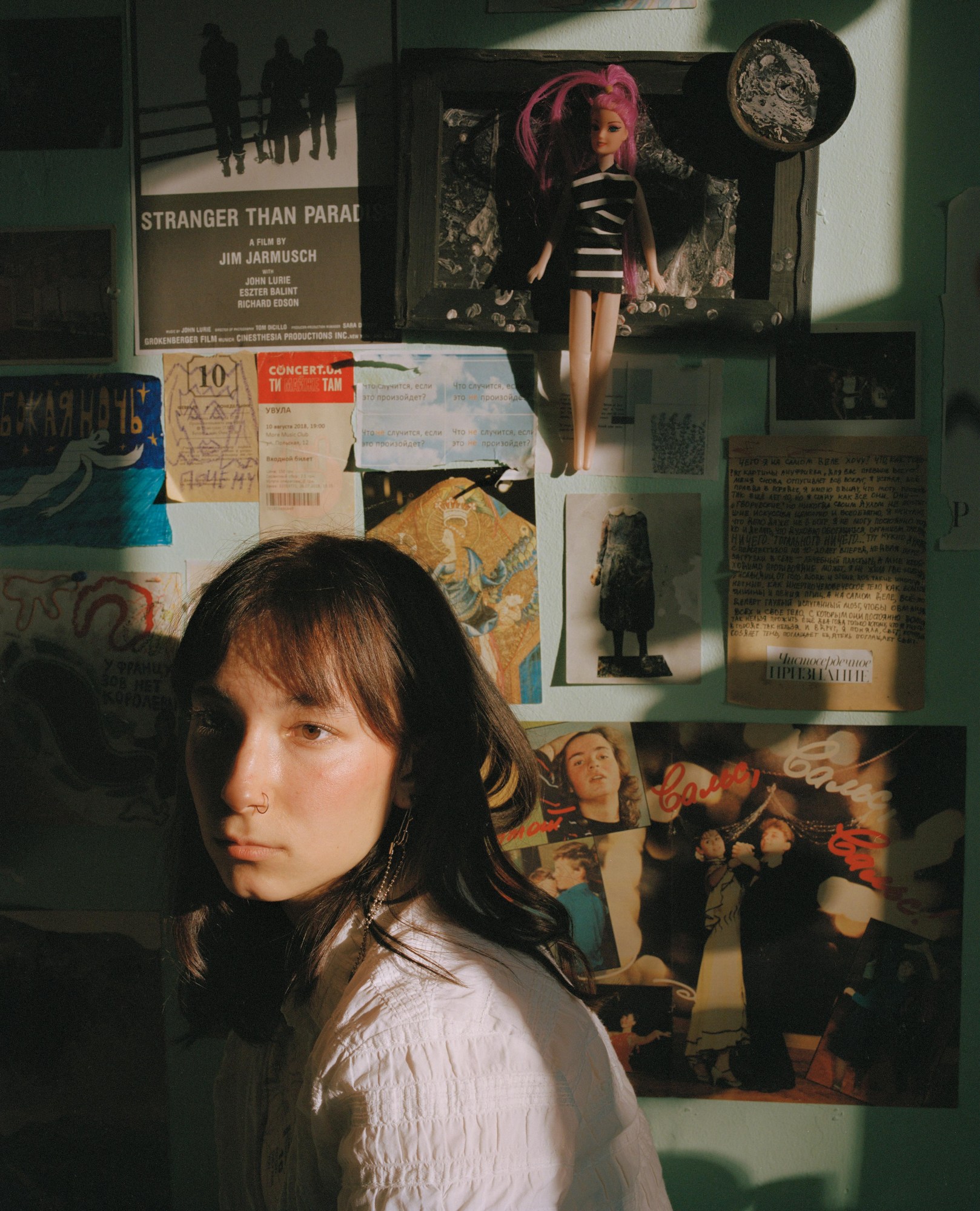 close up of a model in half-sun, the wall behind her covered in pictures, posters and a pink-haired barbie doll