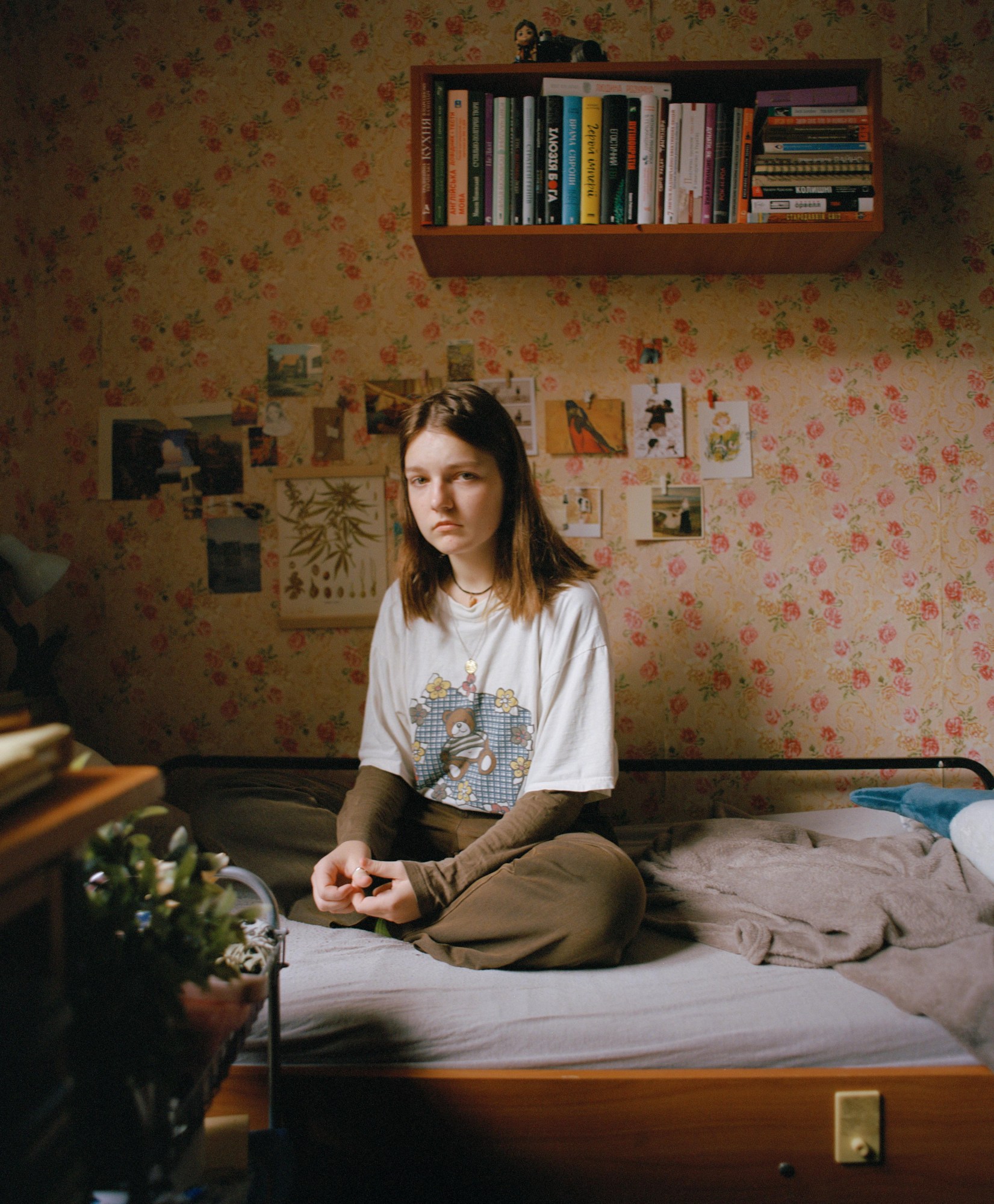 model sits cross legged on a bed facing the camera, yellow and pink flower wallpaper in the background