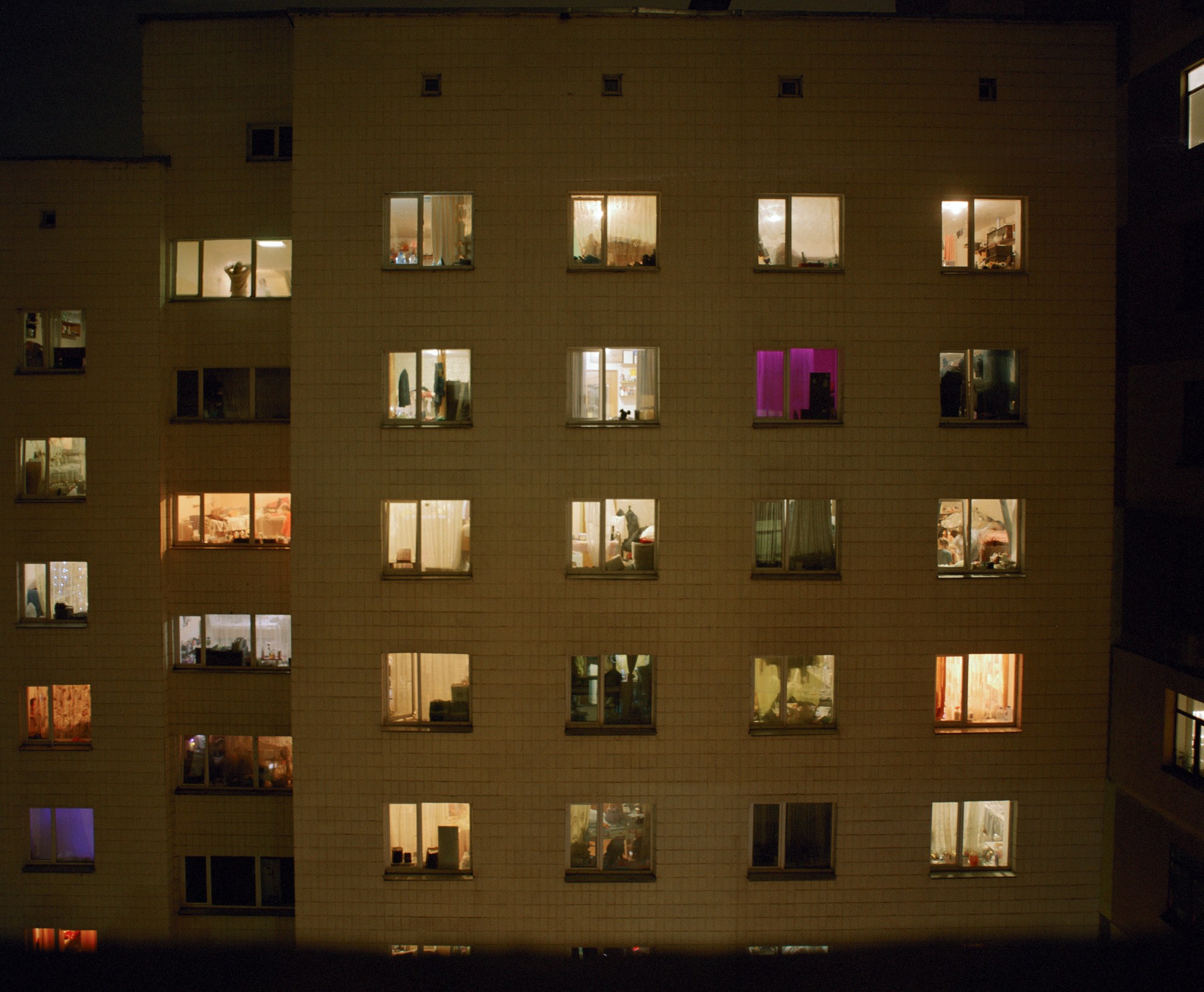 one side of a high rise building, each window slightly different in terms of light (some yellow, some purple) and people (some are silhouetted, some are not visible)