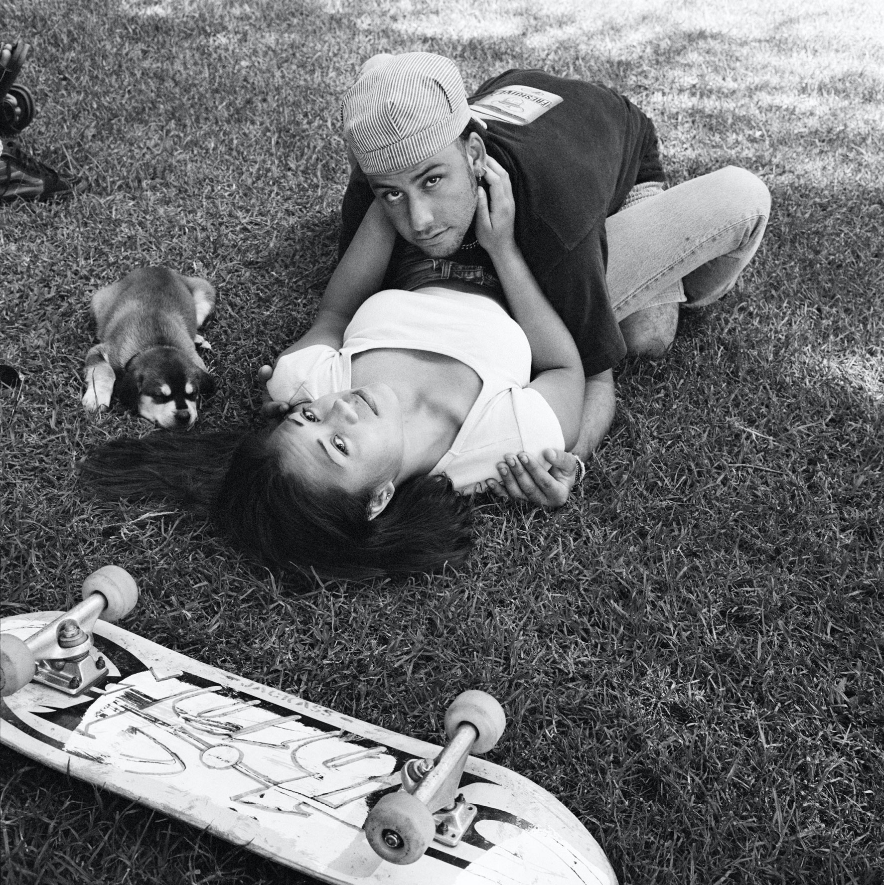 a skateboarder straddles his girlfriend, both looking at the camera, as a puppy sits by them on the grass