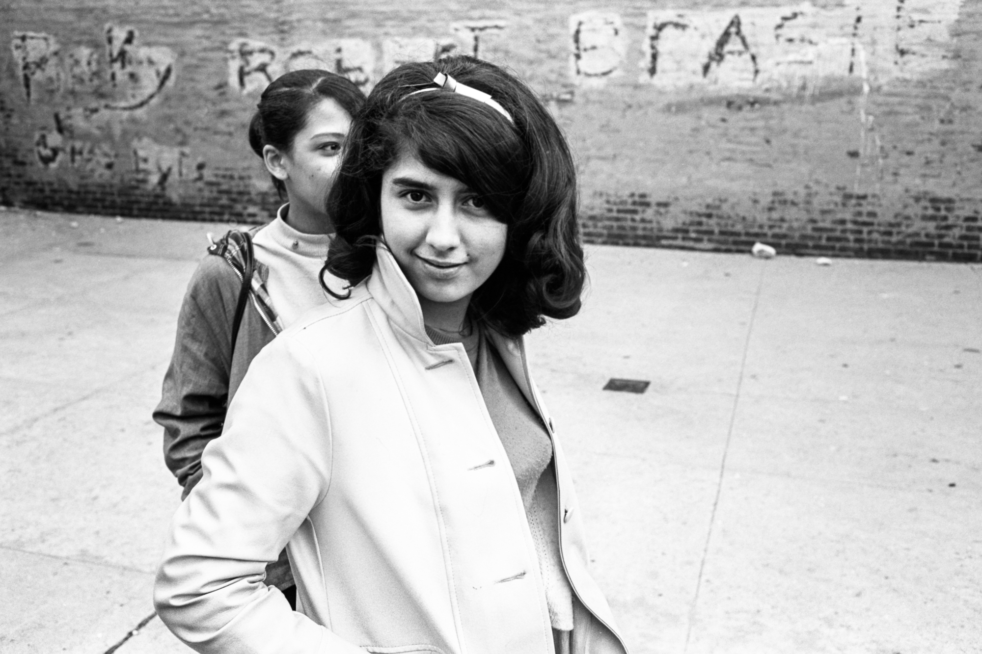 black and white photo of teenage girl in white coat with boy seen behind her