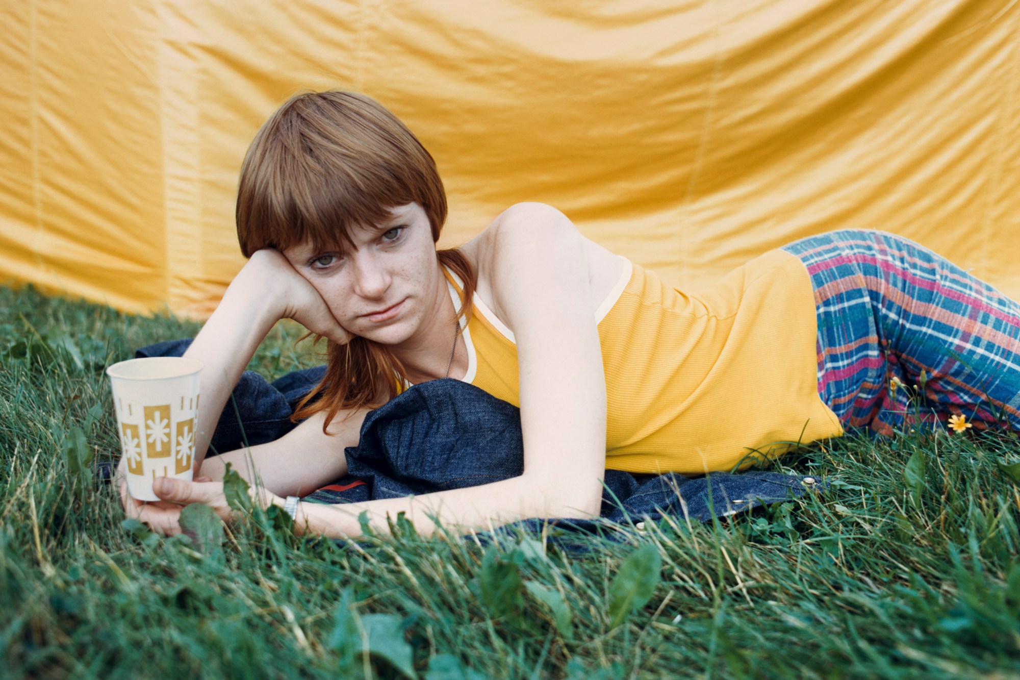 A woman in a yellow top and blue check trousers lies on the grass with a cup in front of a yellow tent.