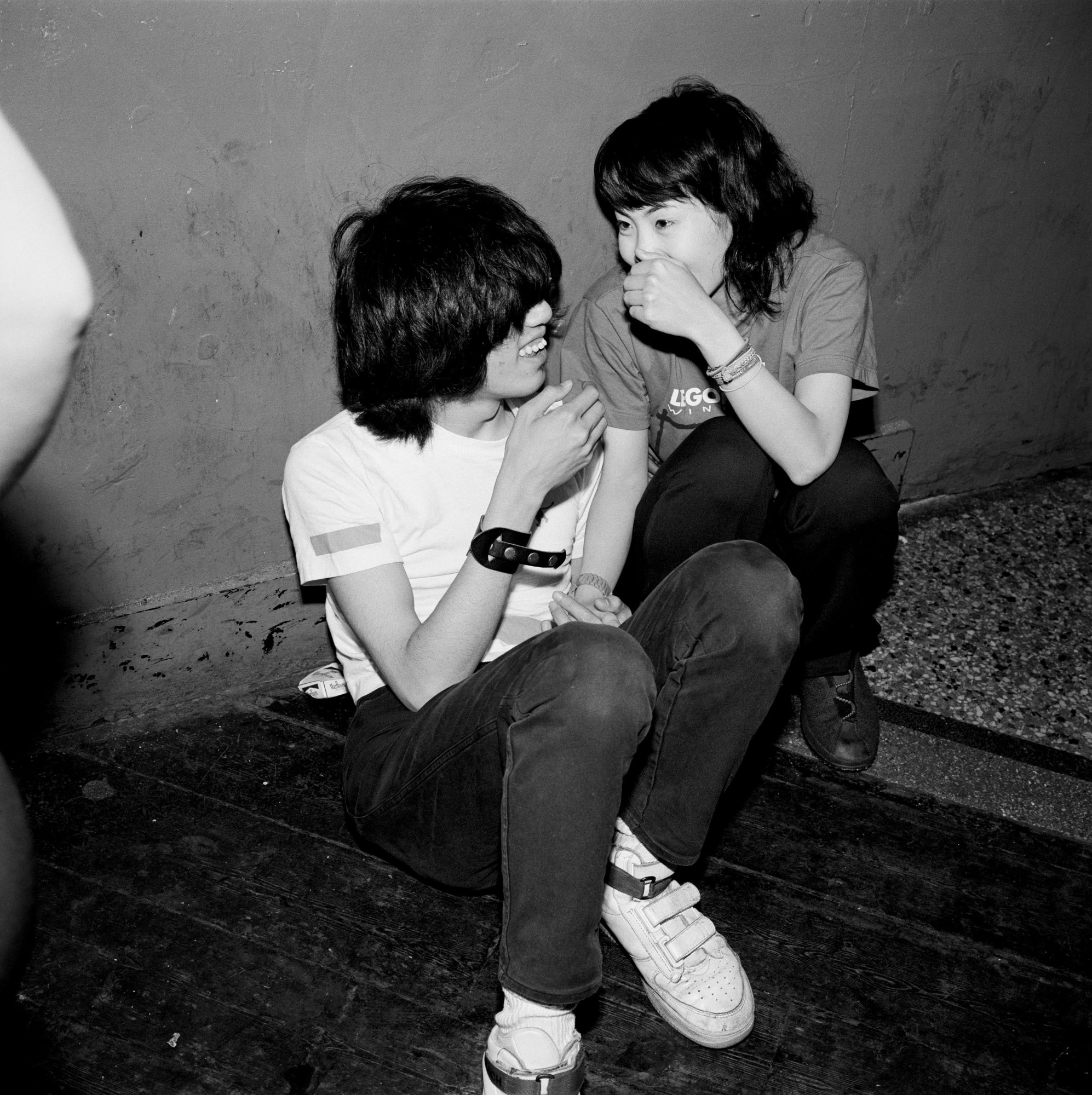 Black and white photo of teenagers whispering to each other while sitting on dancefloor