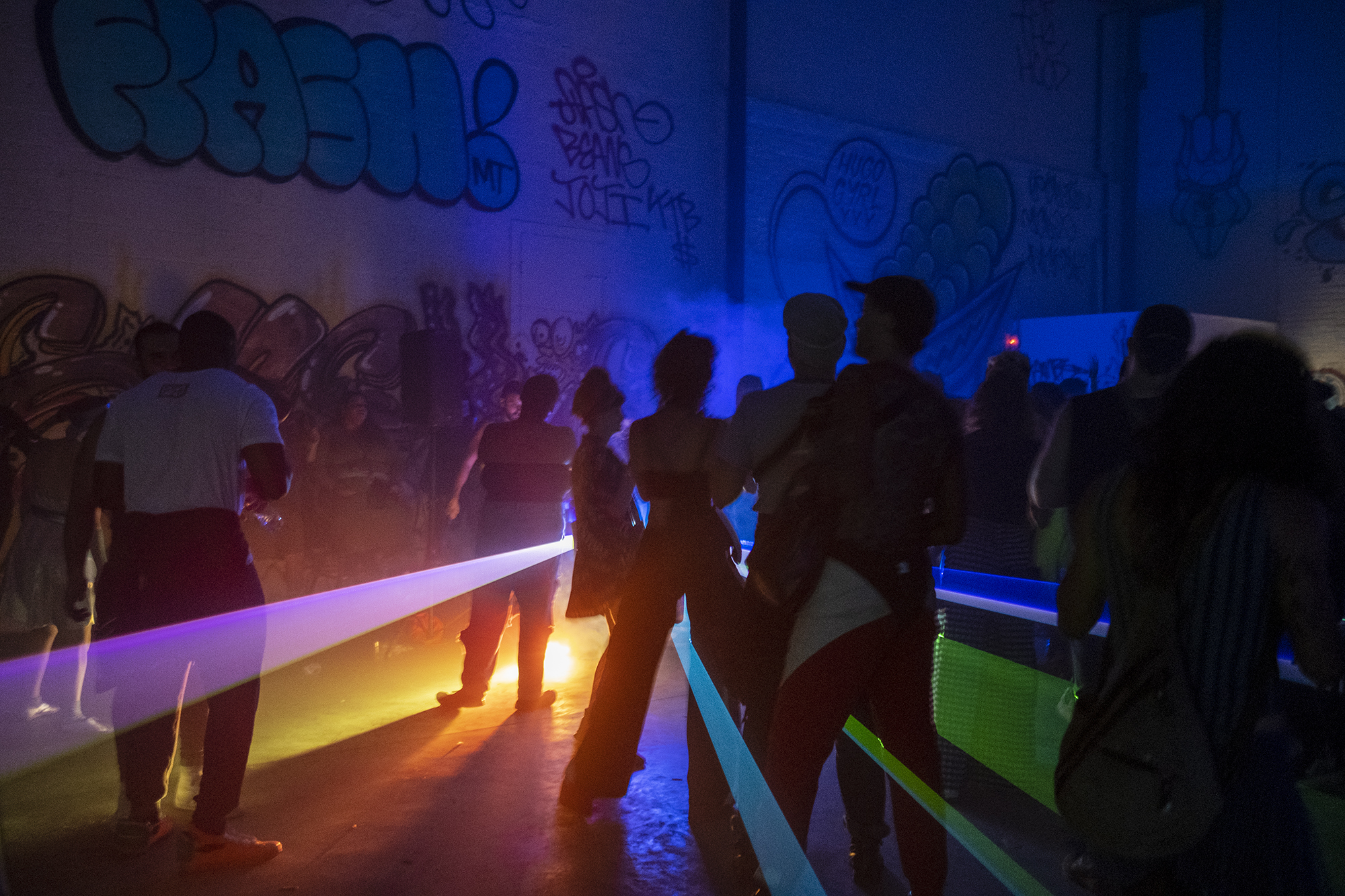 rainbow lasers illuminate a small group of ravers in a warehouse space covered in graffiti