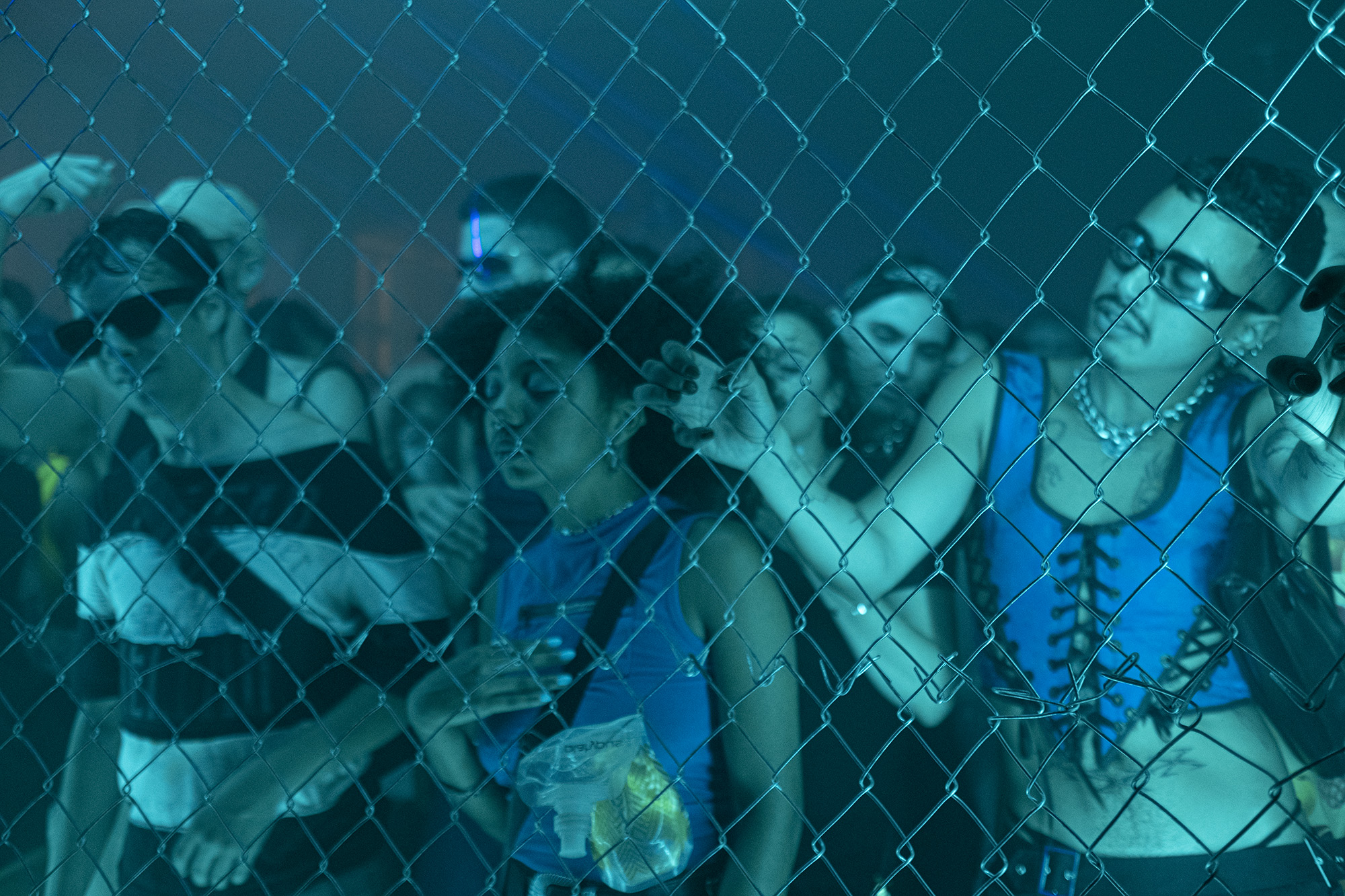 a crowd of club-goers dance behind a chainlink fence under a blue light