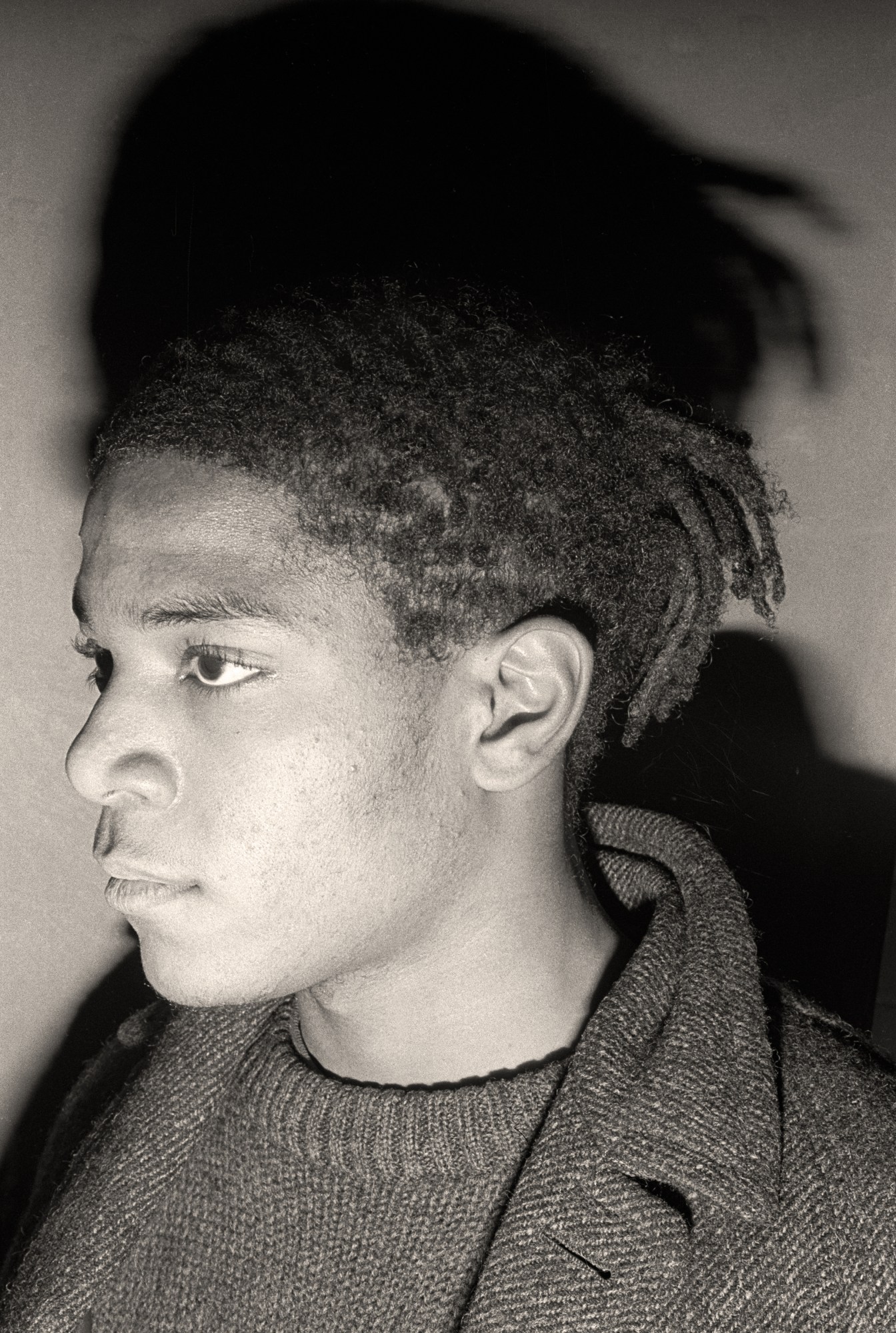 Side portrait of Jean-Michel Basquiat in a jumper and tweed coat photographed by Marcia Resnick.