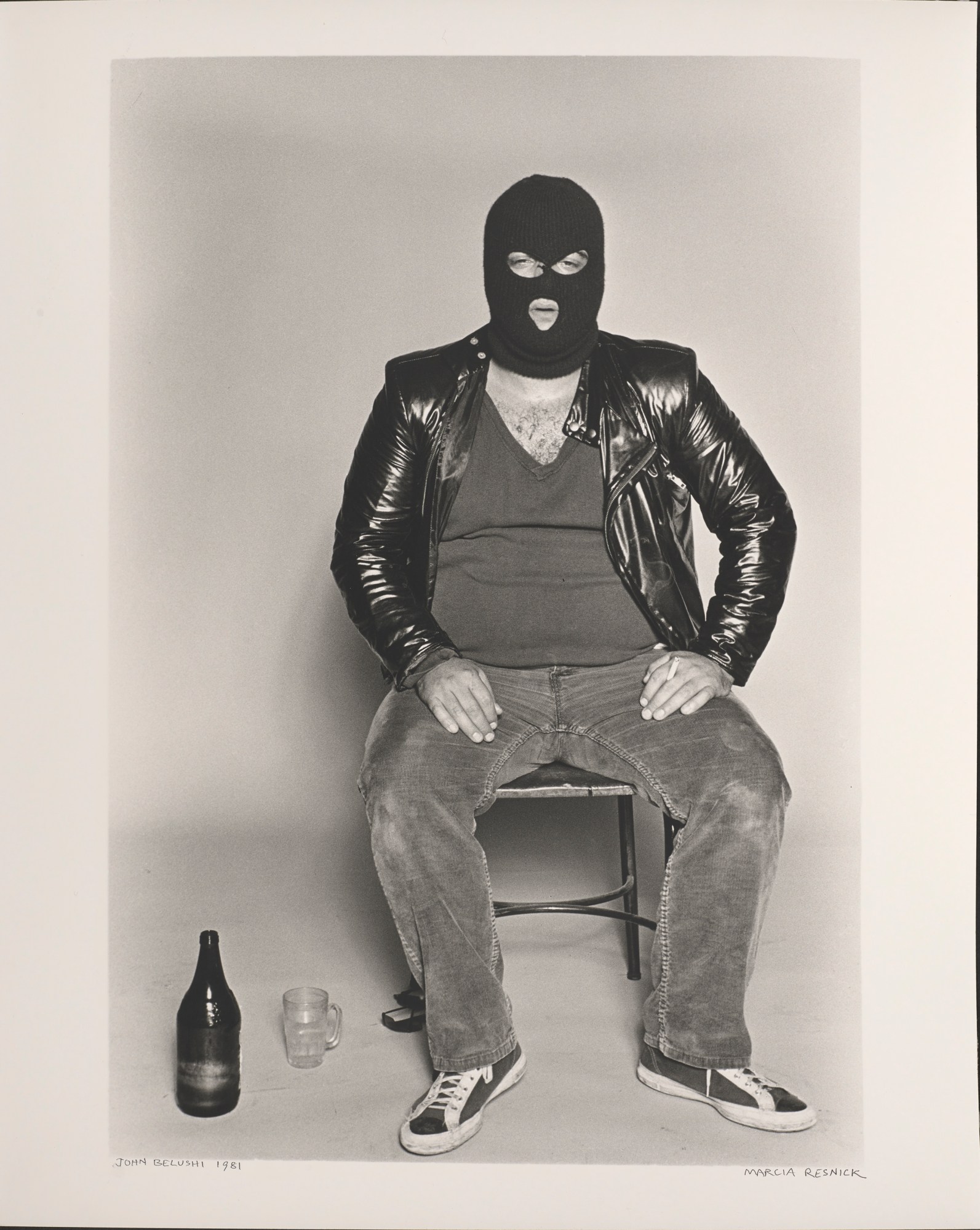 Portrait of John Belushi sitting down on a chair in a vest, jeans, trainers, latex jacket and face mask with a drink and glass next to him photographed by Marcia Resnick.