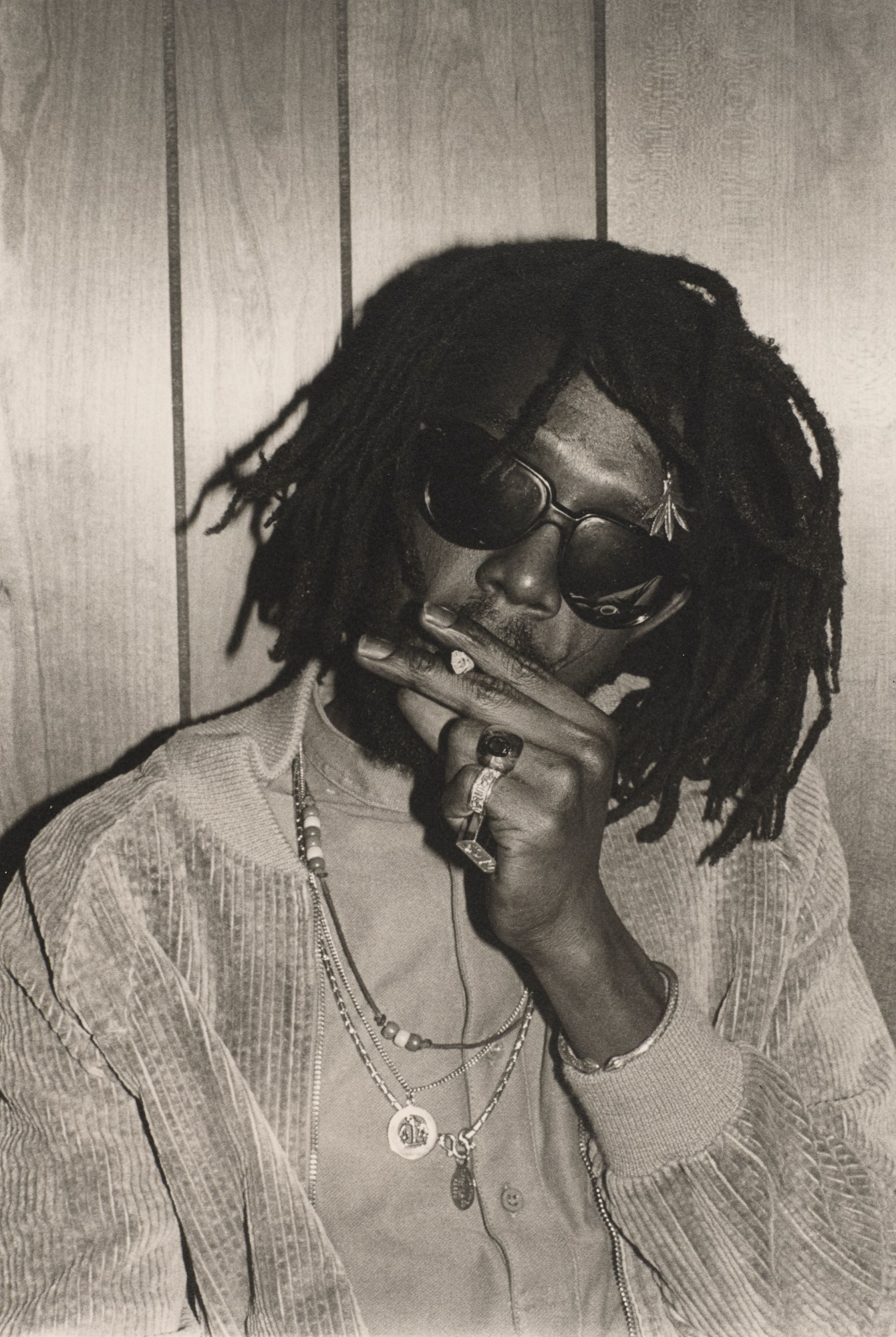 Photograph of Peter Tosh in a jacket, collarless shirt and necklaces smoking by Marcia Resnick.