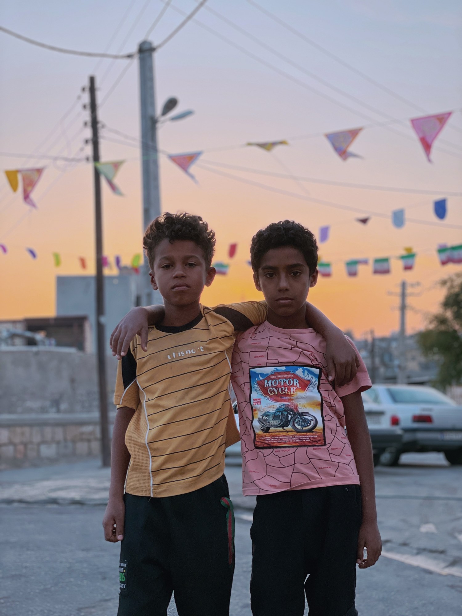 Two young boys with their arms around each other standing on the street at sunset.