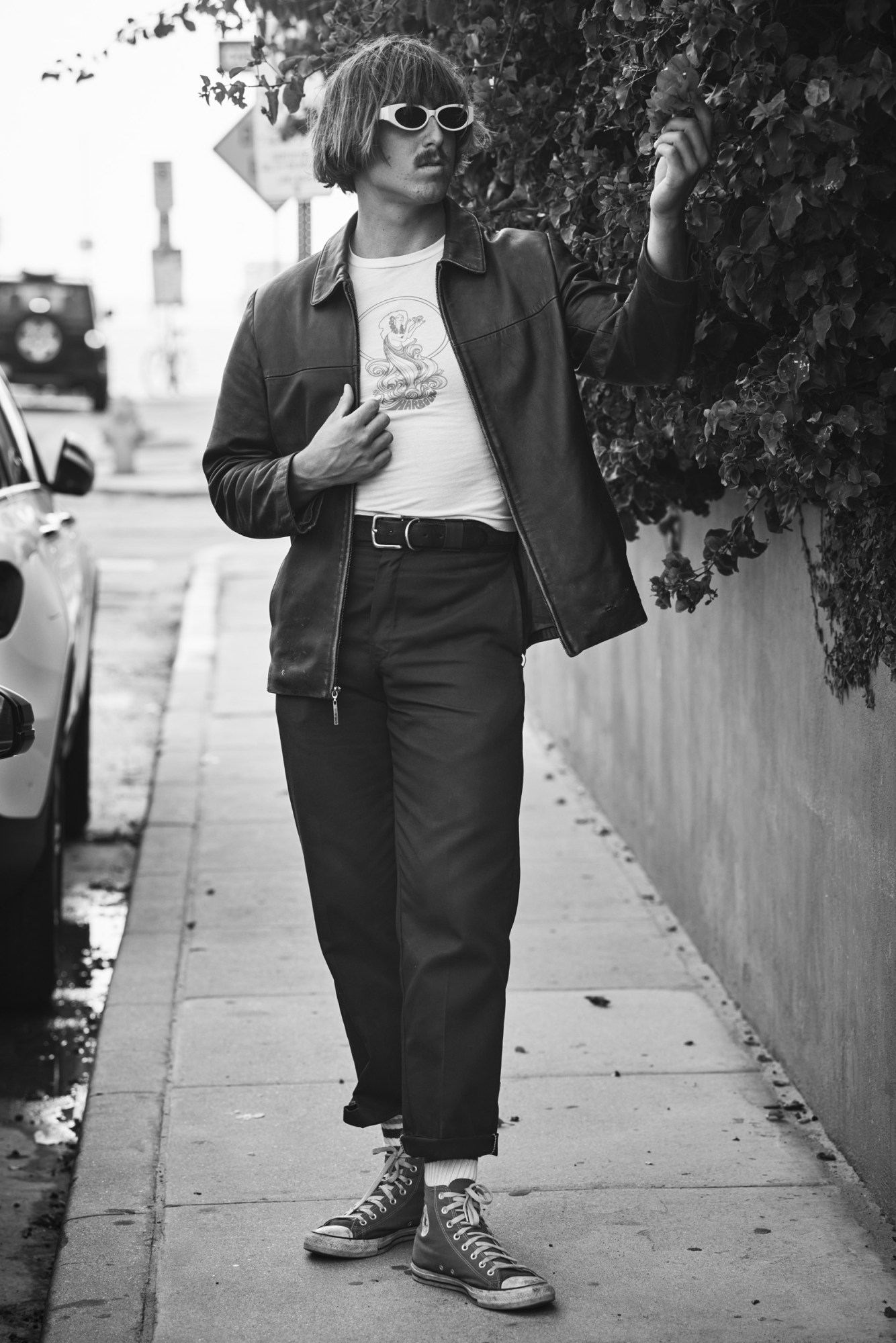 A man wearing converse and sunglasses looking at a flower.