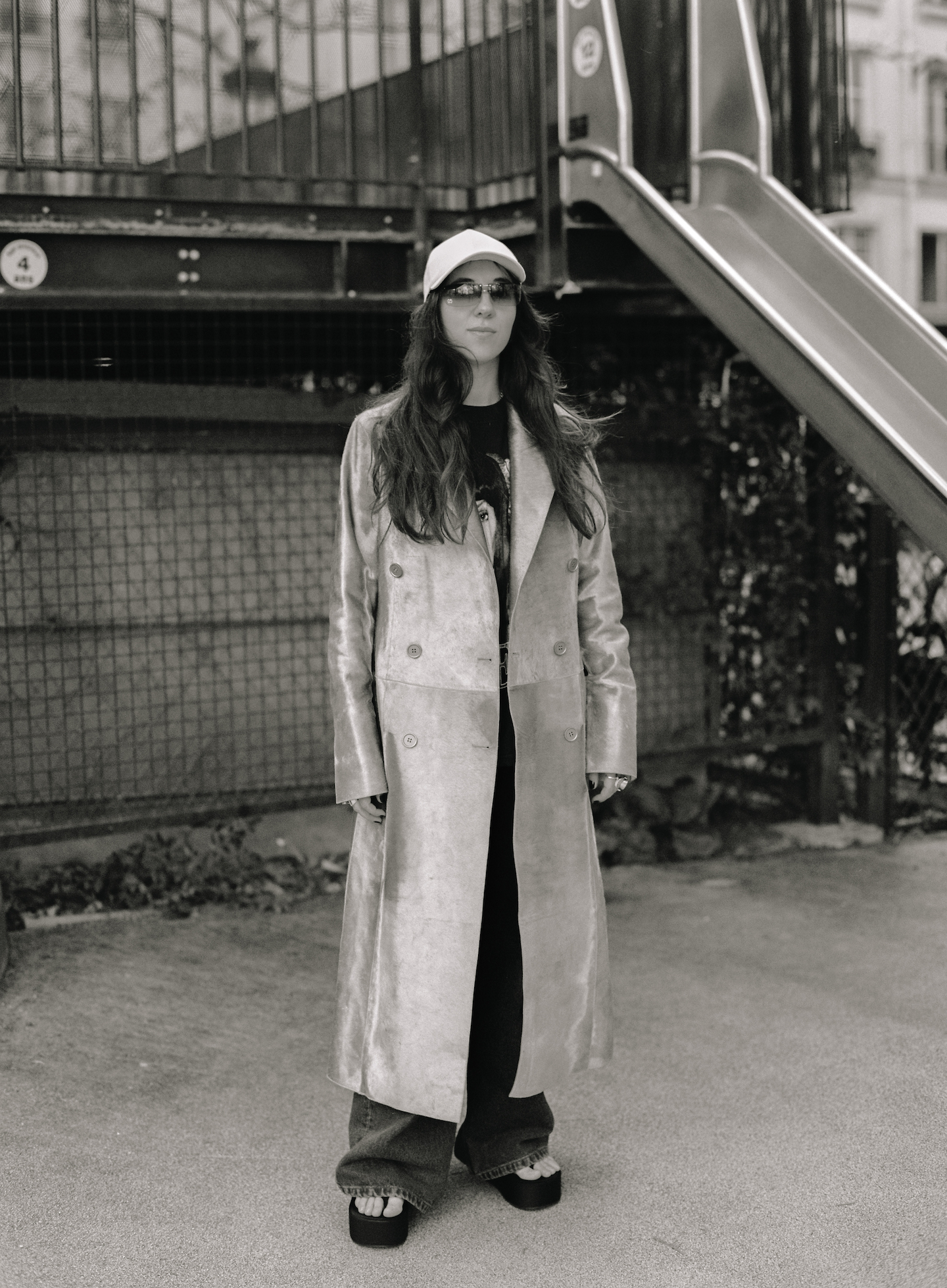 Chloe Caillet wearing a ling leather coat, cap and sunglasses, standing by a slide