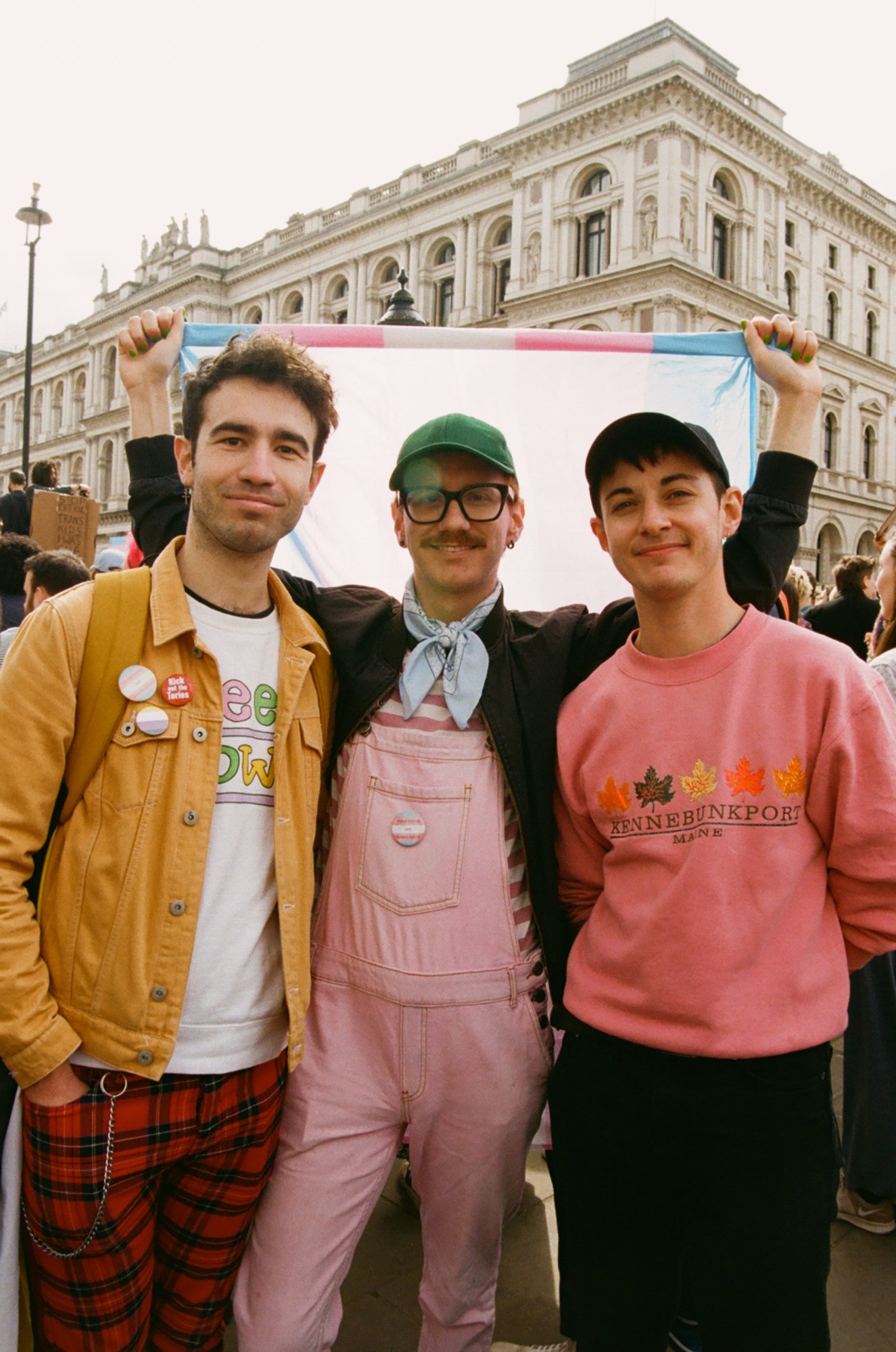 Three protesters standing under a trans flag at the ban trans conversion therapy protest at Downing Street, London.