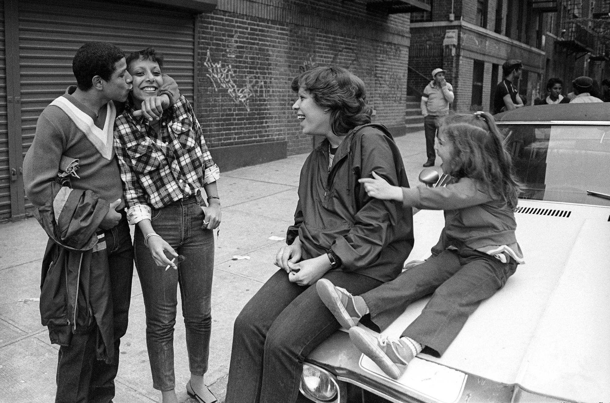 a couple embrace in front of a pair of kids lounging on the hood of a car