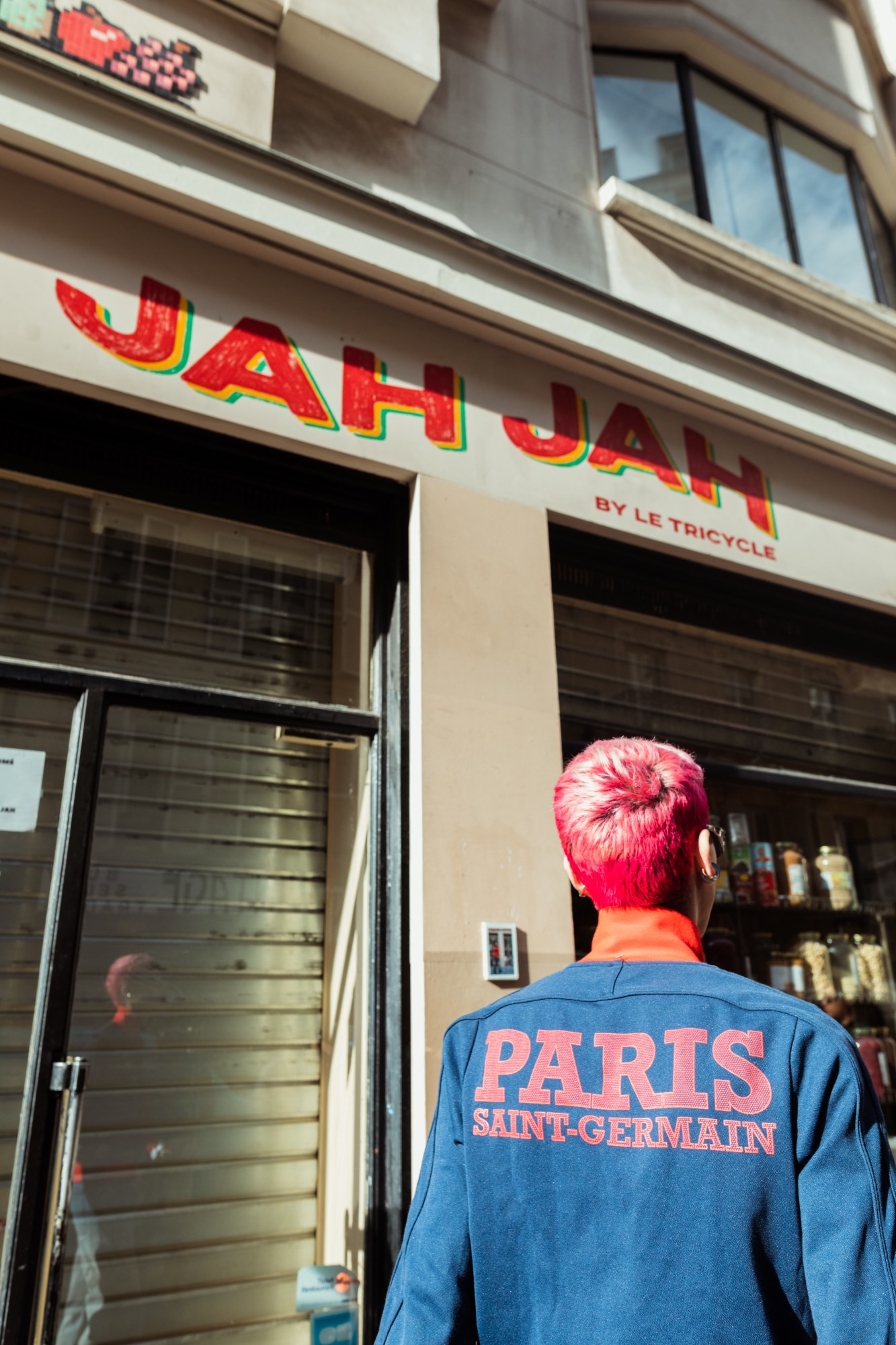 a person with cropped bright pink hair stands in front of the Jah Jah cafe sign in Paris