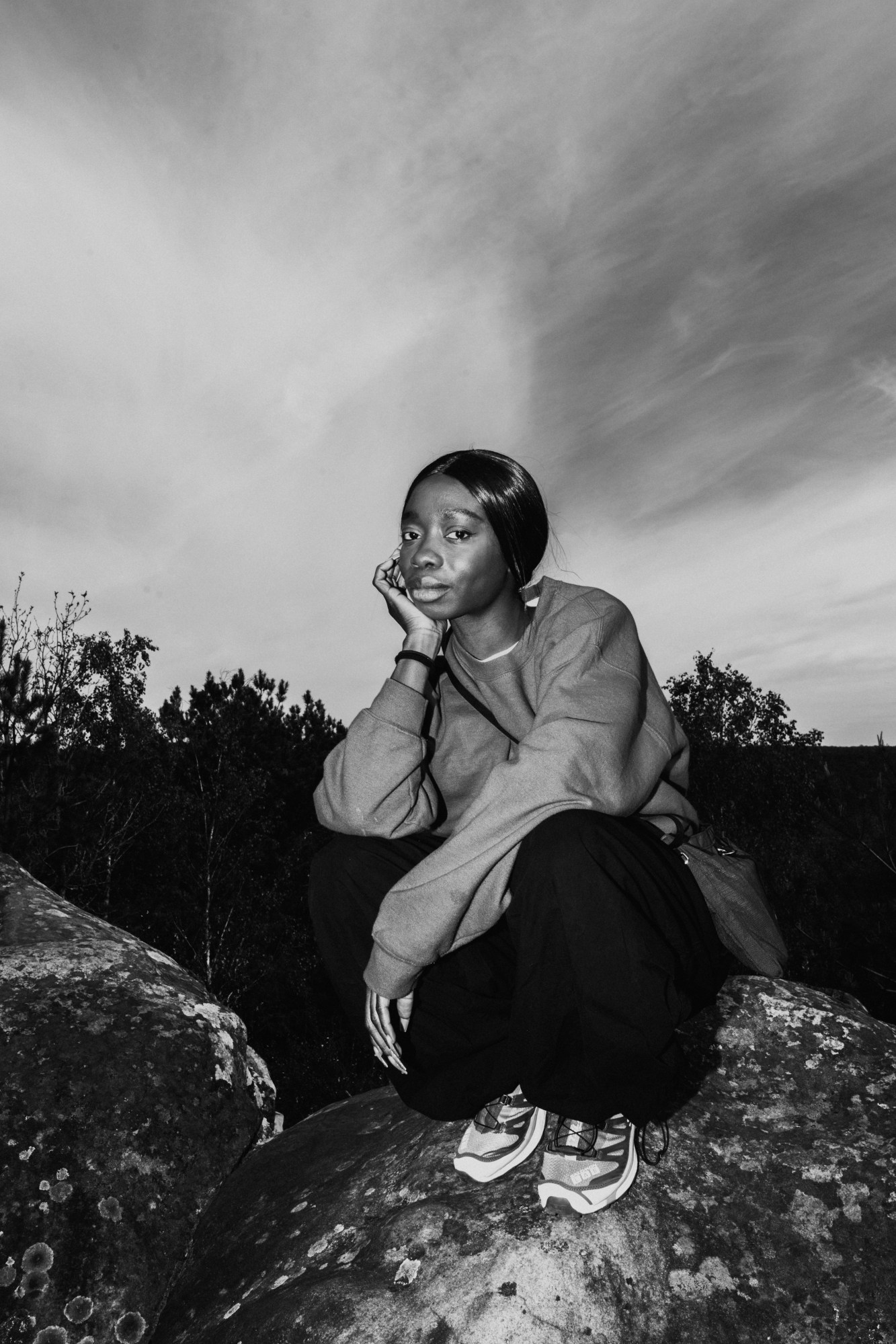 a young woman squats on a rock in nature, cradling her face and looking to the camera