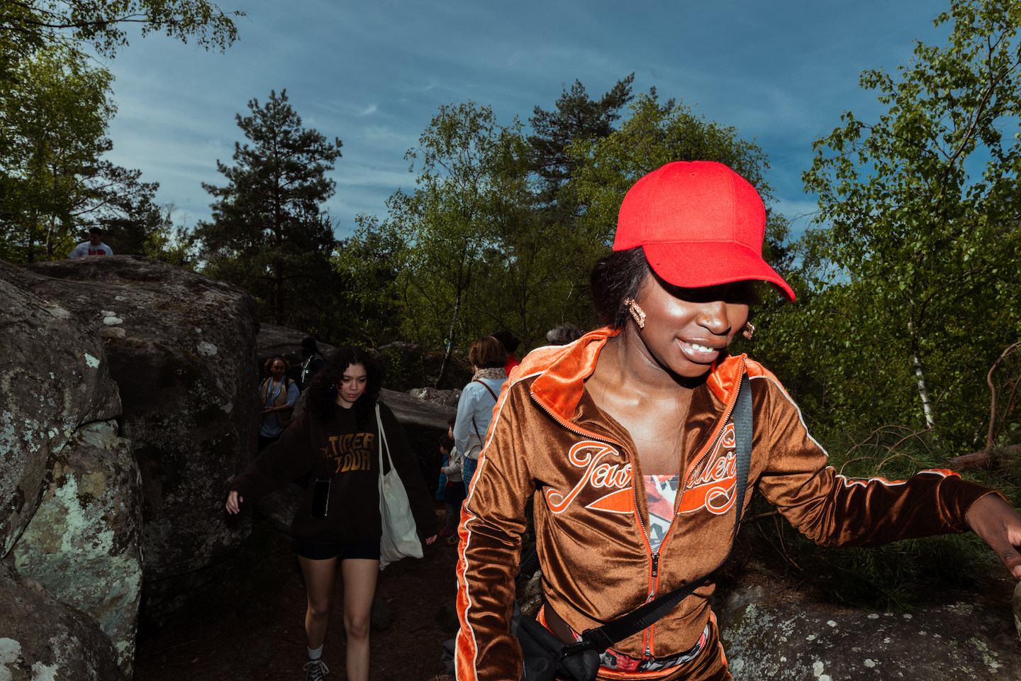 a woman in a brown velour tracksuit and red cap is photographed with flash as she hikes in a forest