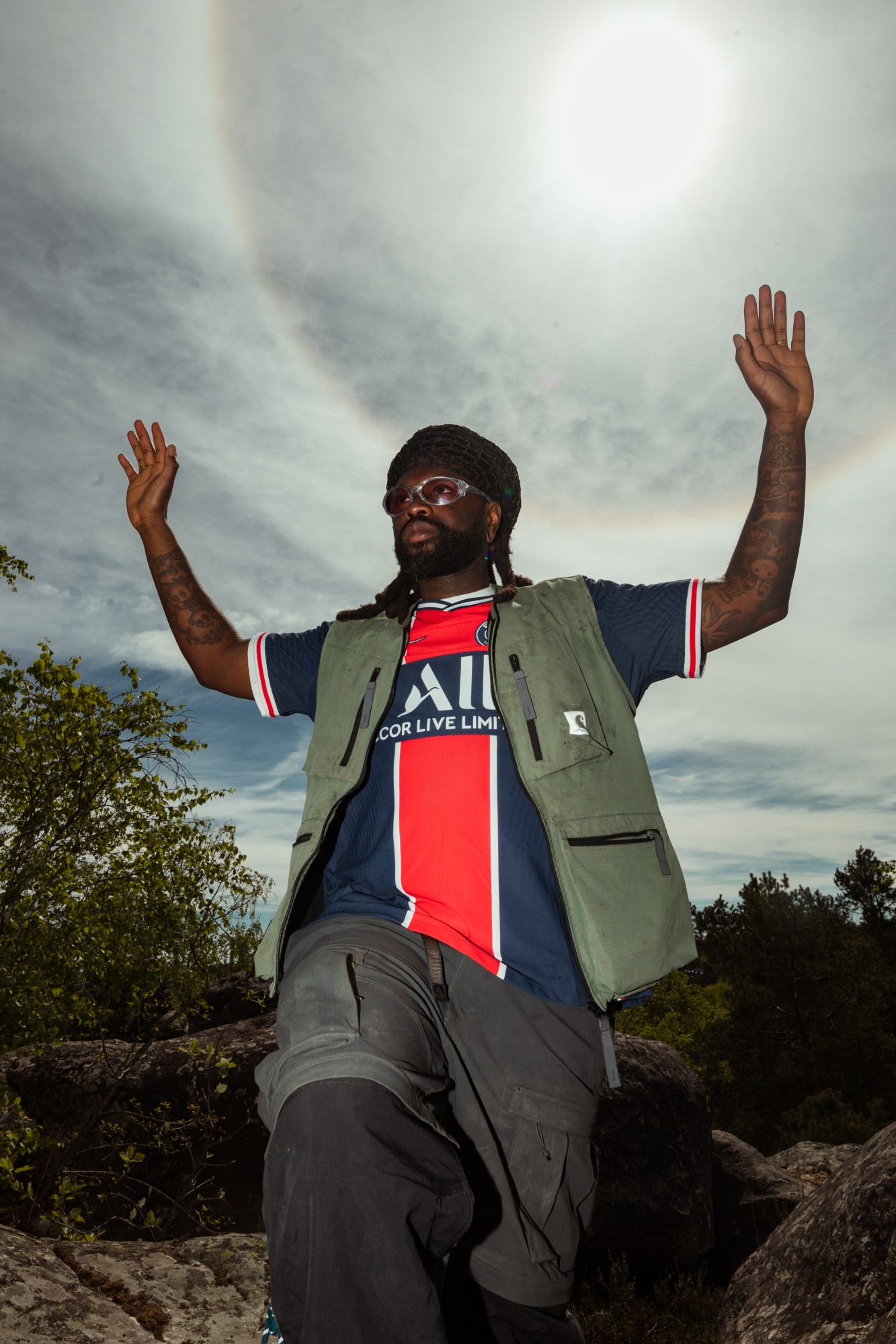 a man with dreads wearing 90s sunglasses and a hat stands in nature, his arms raised above his head