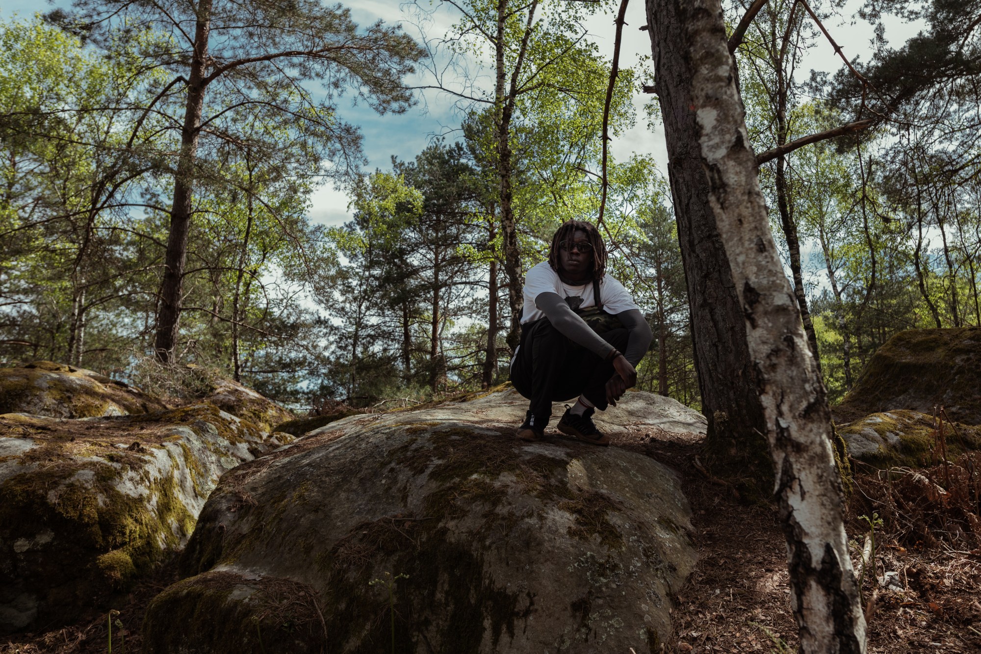 a person squats on a rock by a tree, looking to camera, surrounded by forest