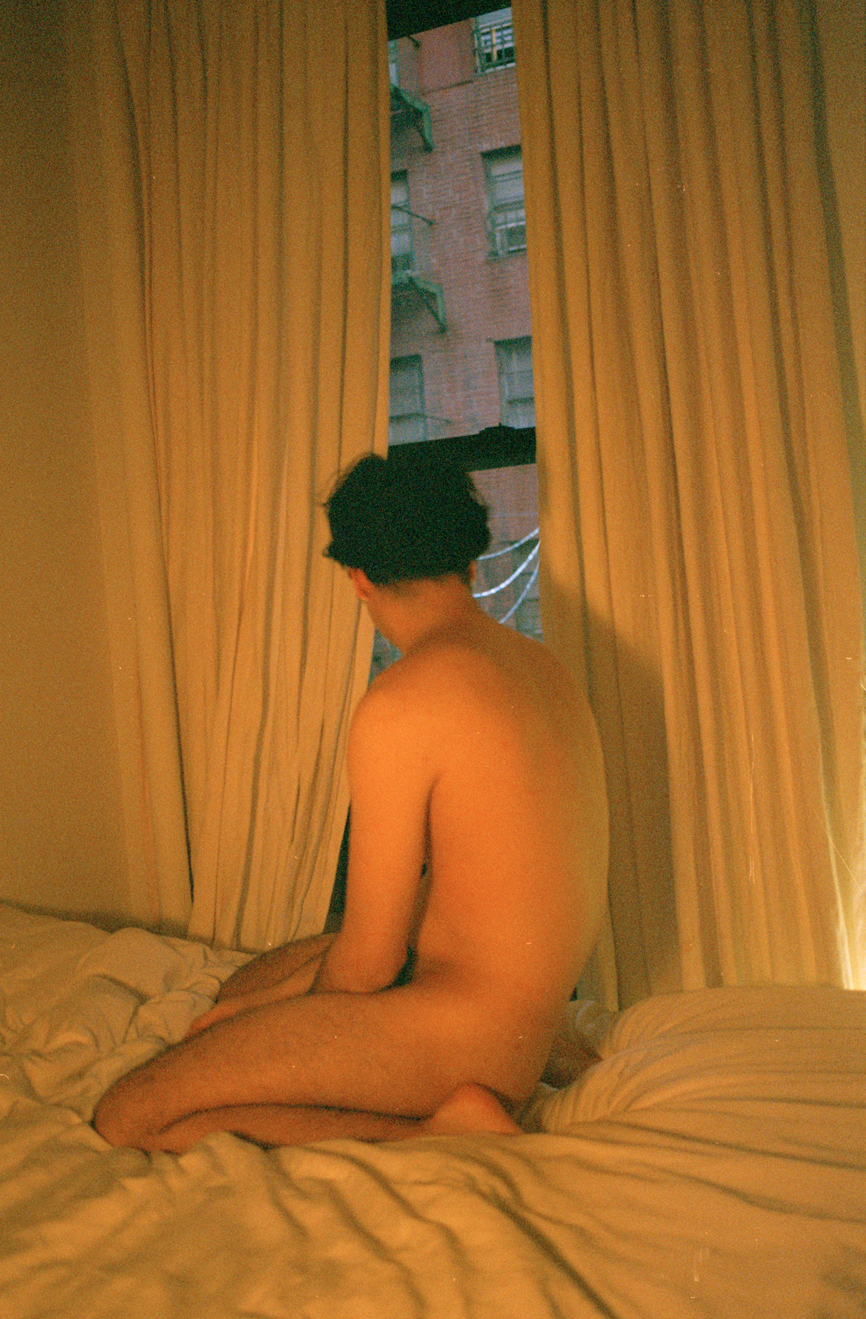 a naked person in a room, bathed in yellow light, peering through curtains