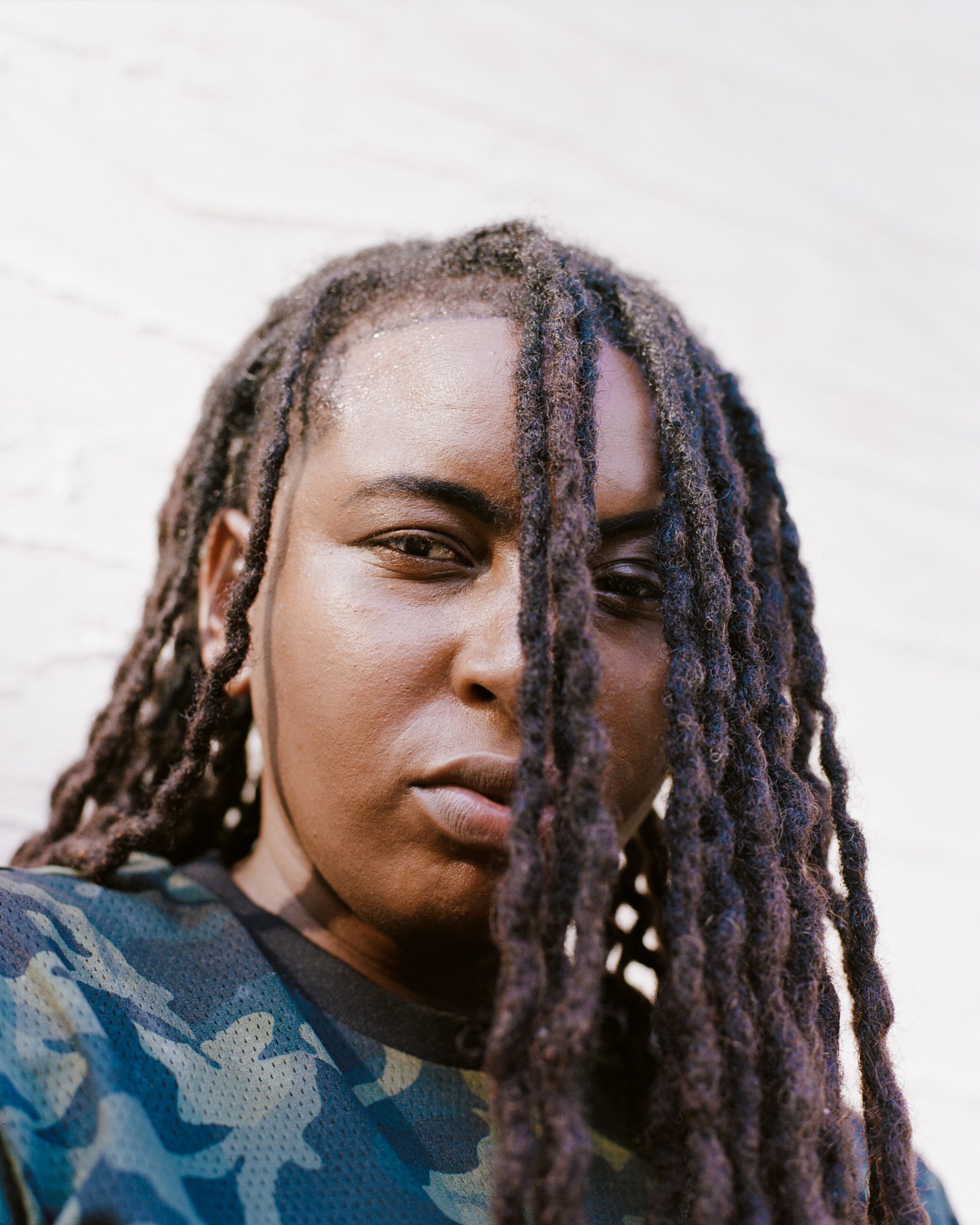 a model wearing a camouflage print shirt looks at the camera, their face obscured by locs