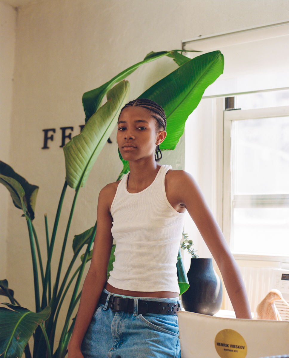 a model dressed in a white vest and blue jeans poses in front of a large house plant