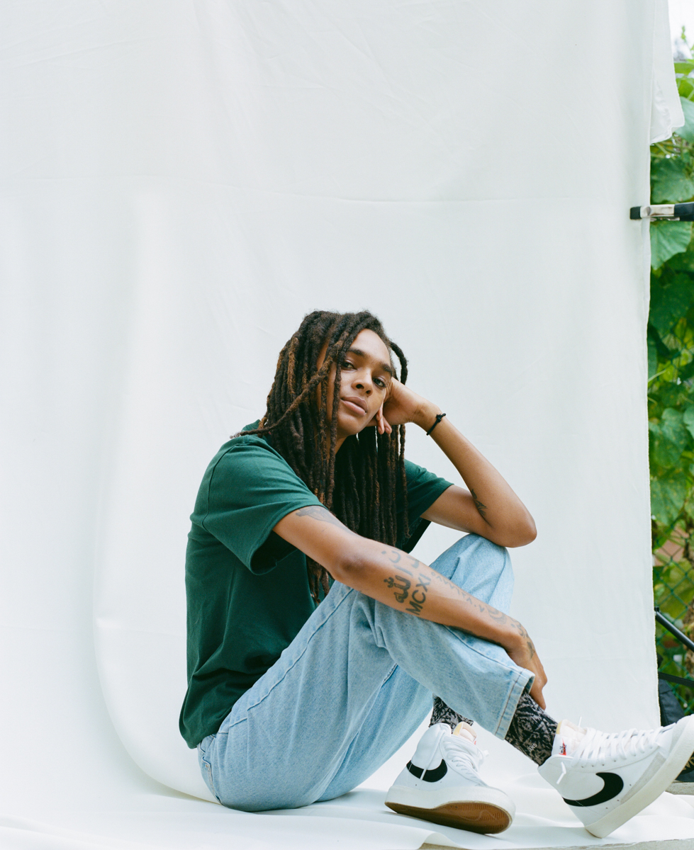 a model is seated on a white backdrop in a green space, wearing pale blue jeans and a dark green t shirt