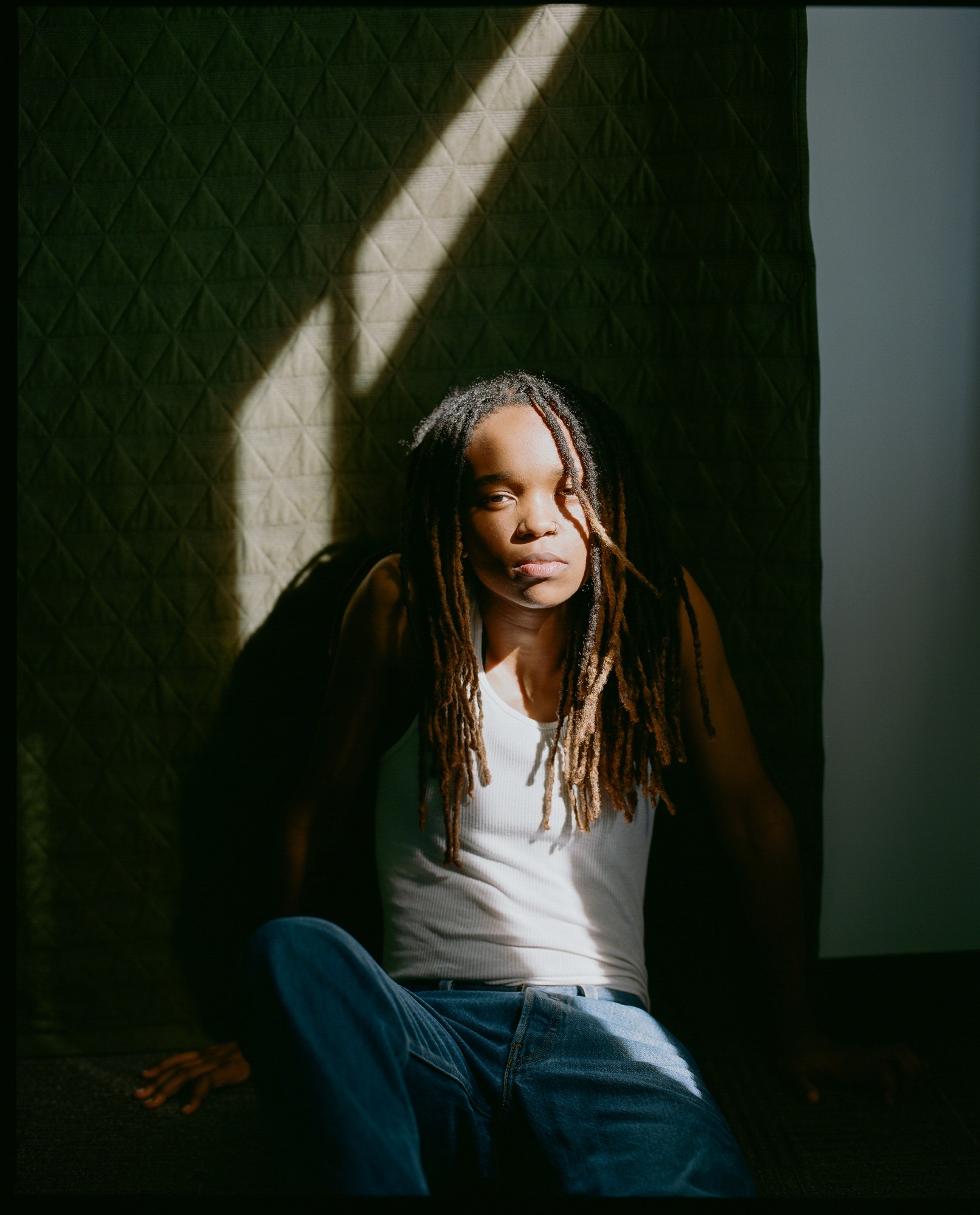 a model in a white vest and jeans sits on the floor in dappled sunlight