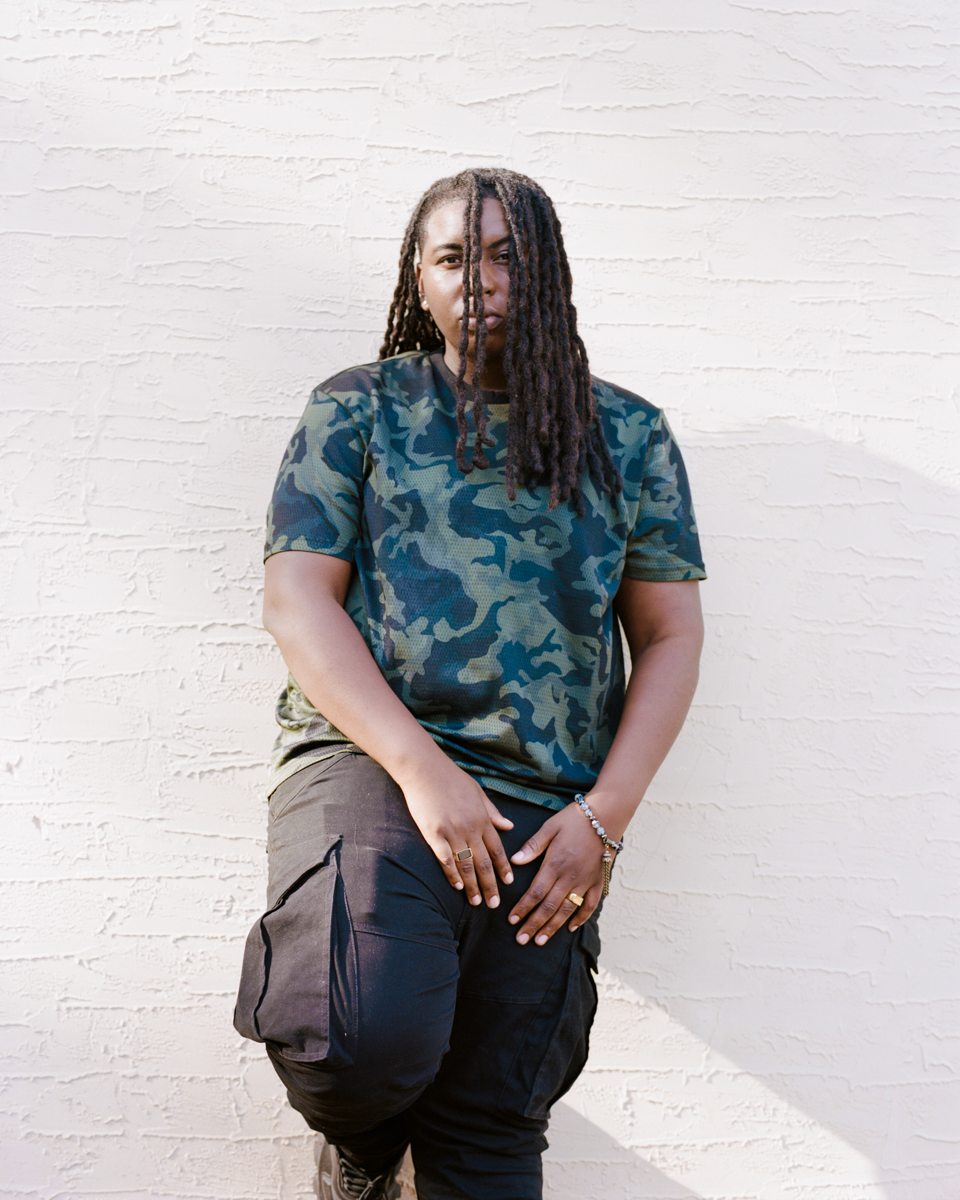 a model leans on a white wall, wearing a camo print shirt and cargo pants and looking to the camera through their hair