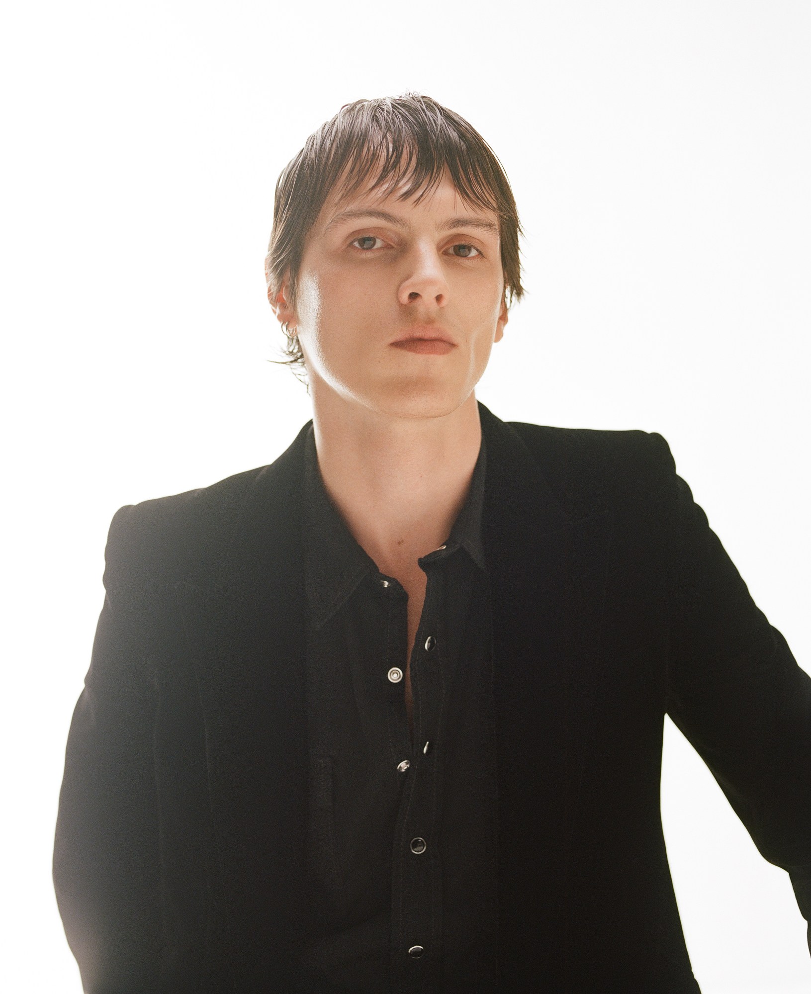 a clean-shaven member of the band Keep Dancing Inc, in a black shirt and suit jacket, looking to camera