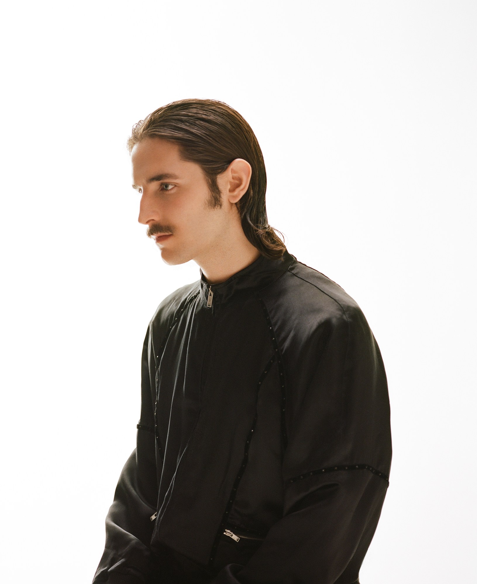 a member of the band Keep Dancing Inc, in a black jacket with slicked back hair and a moustache