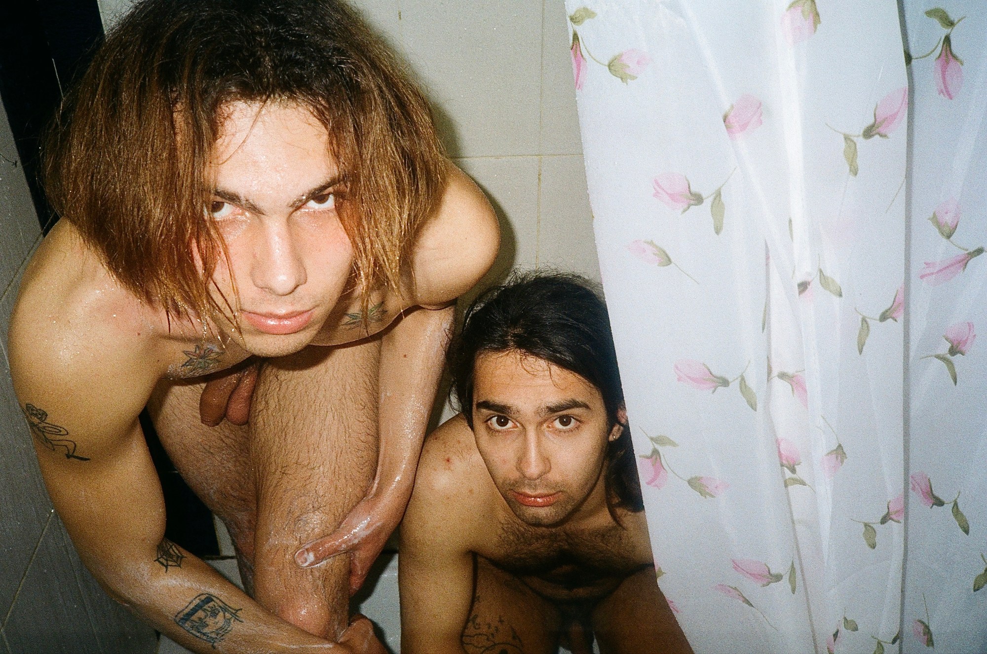 Two men in the shower standing behind a floral shower curtain.