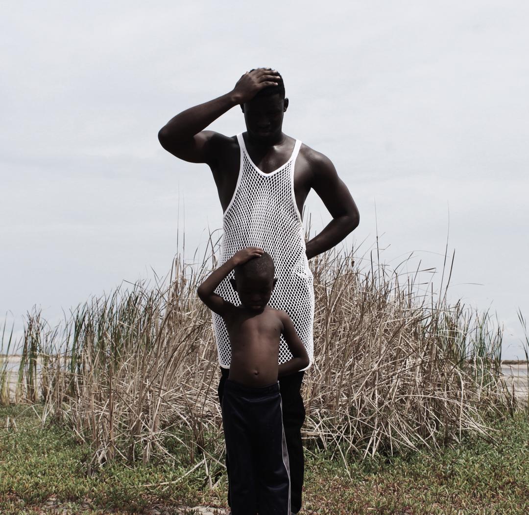 A father and son posing near the ocean, each with a hand on top of his head.