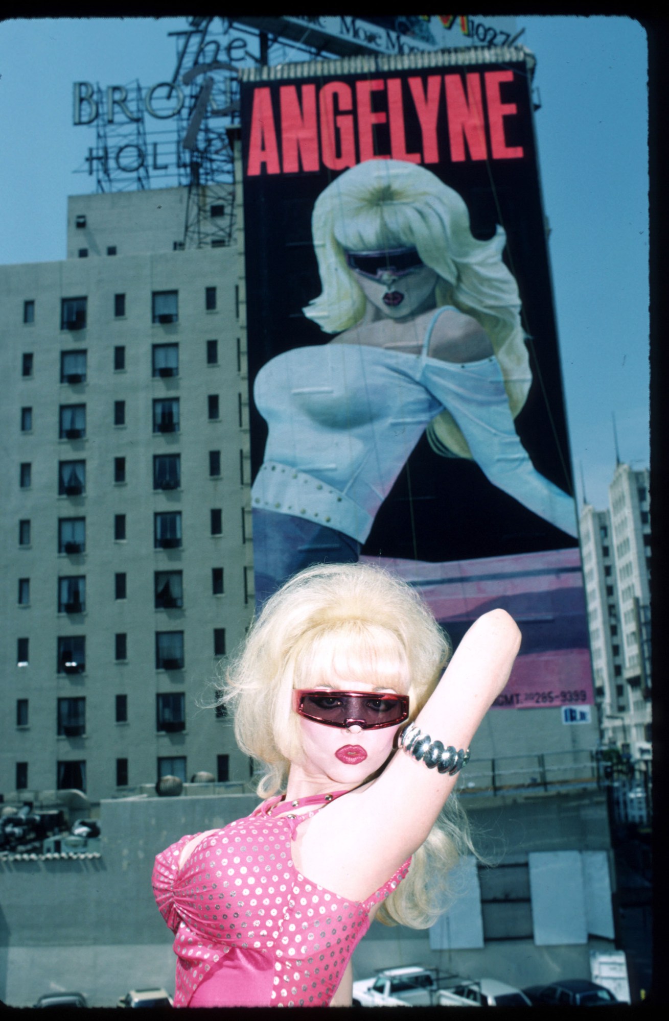 angelyne posing in front of an LA billboard of herself wearing sunglasses in 1987