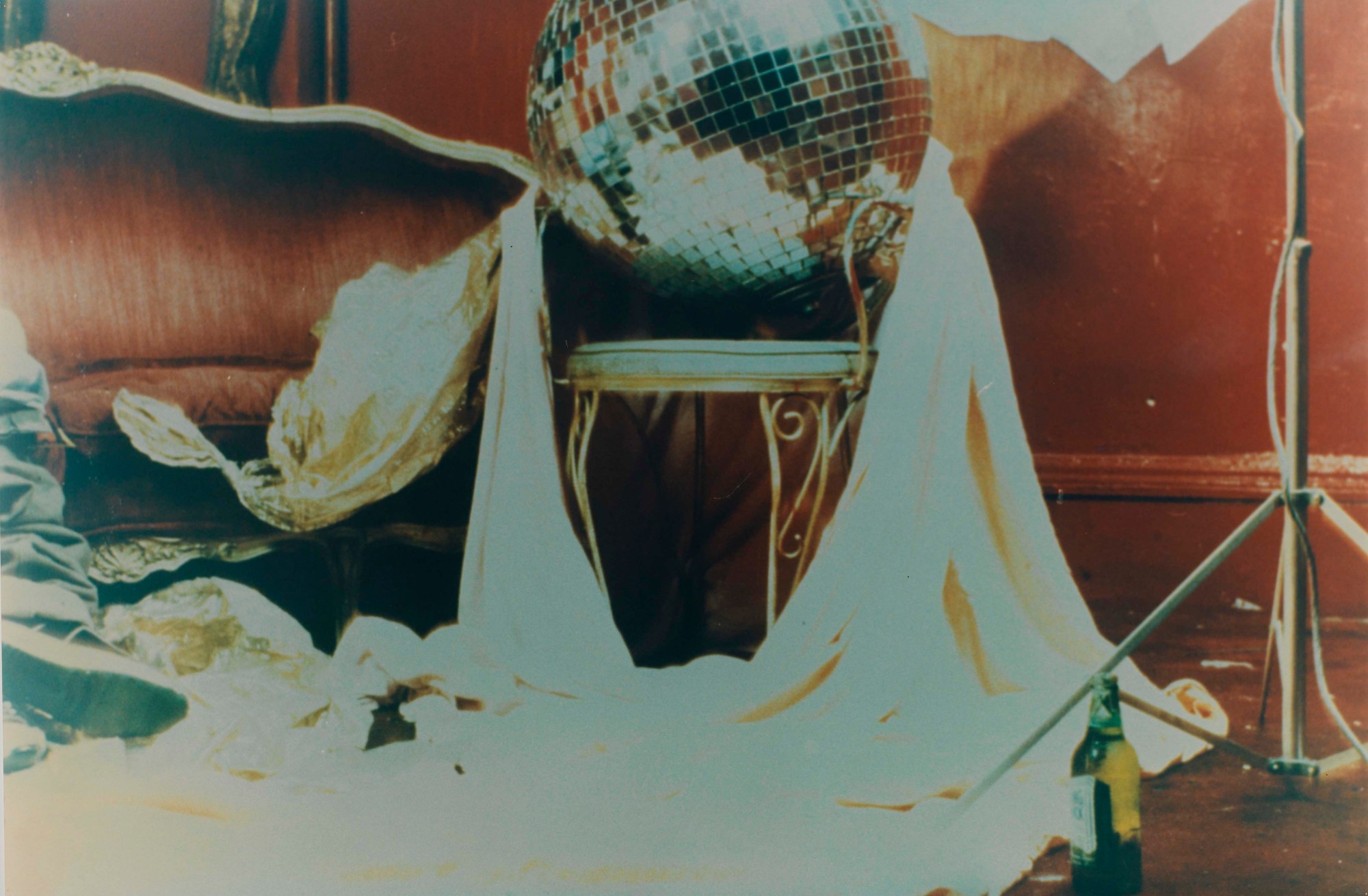 A disco ball sitting on a chair with a sheet around it, a beer and a pair of jeans and trainers.