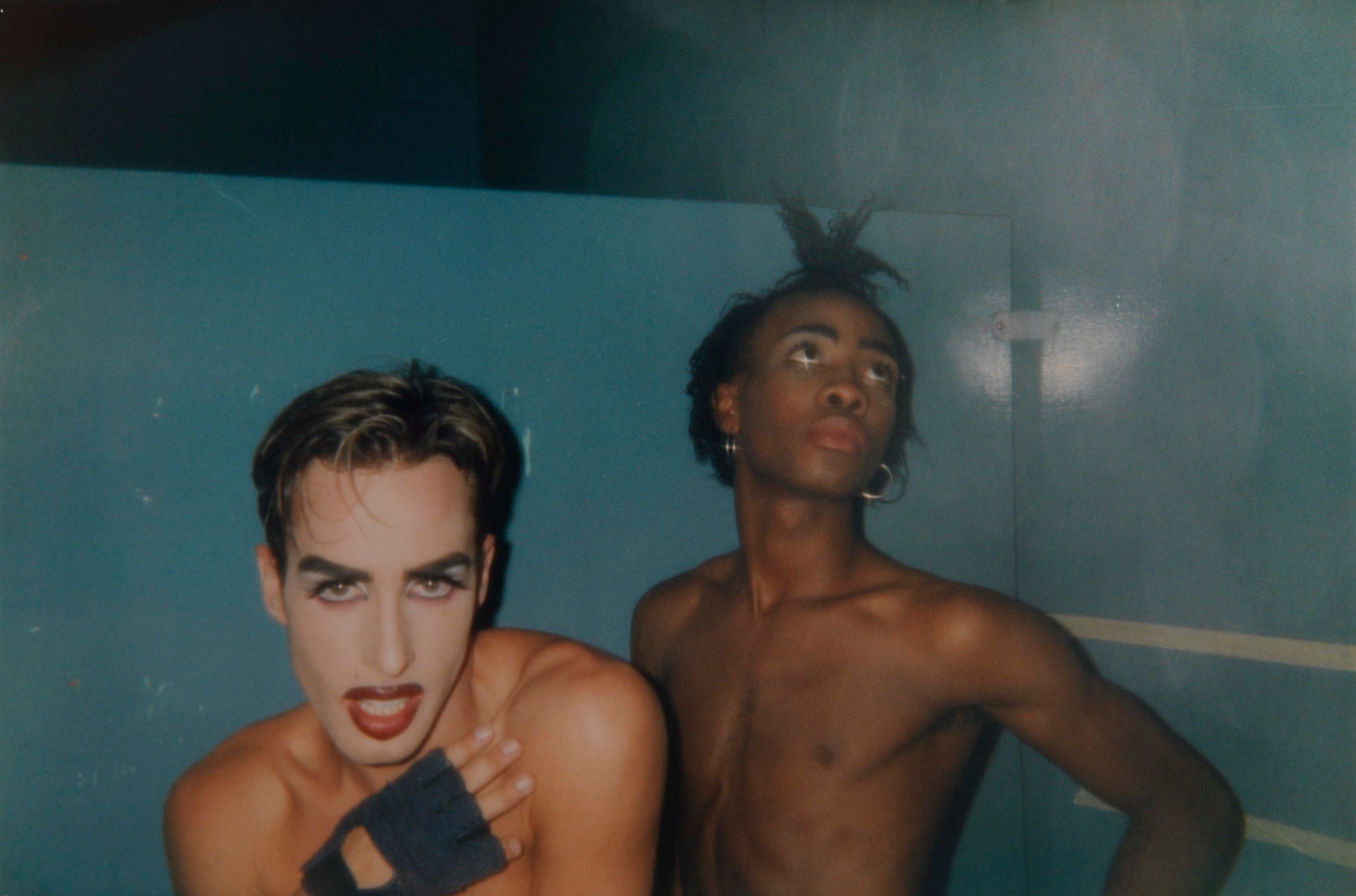 Two people wearing makeup while posing in a mirror shirtless.