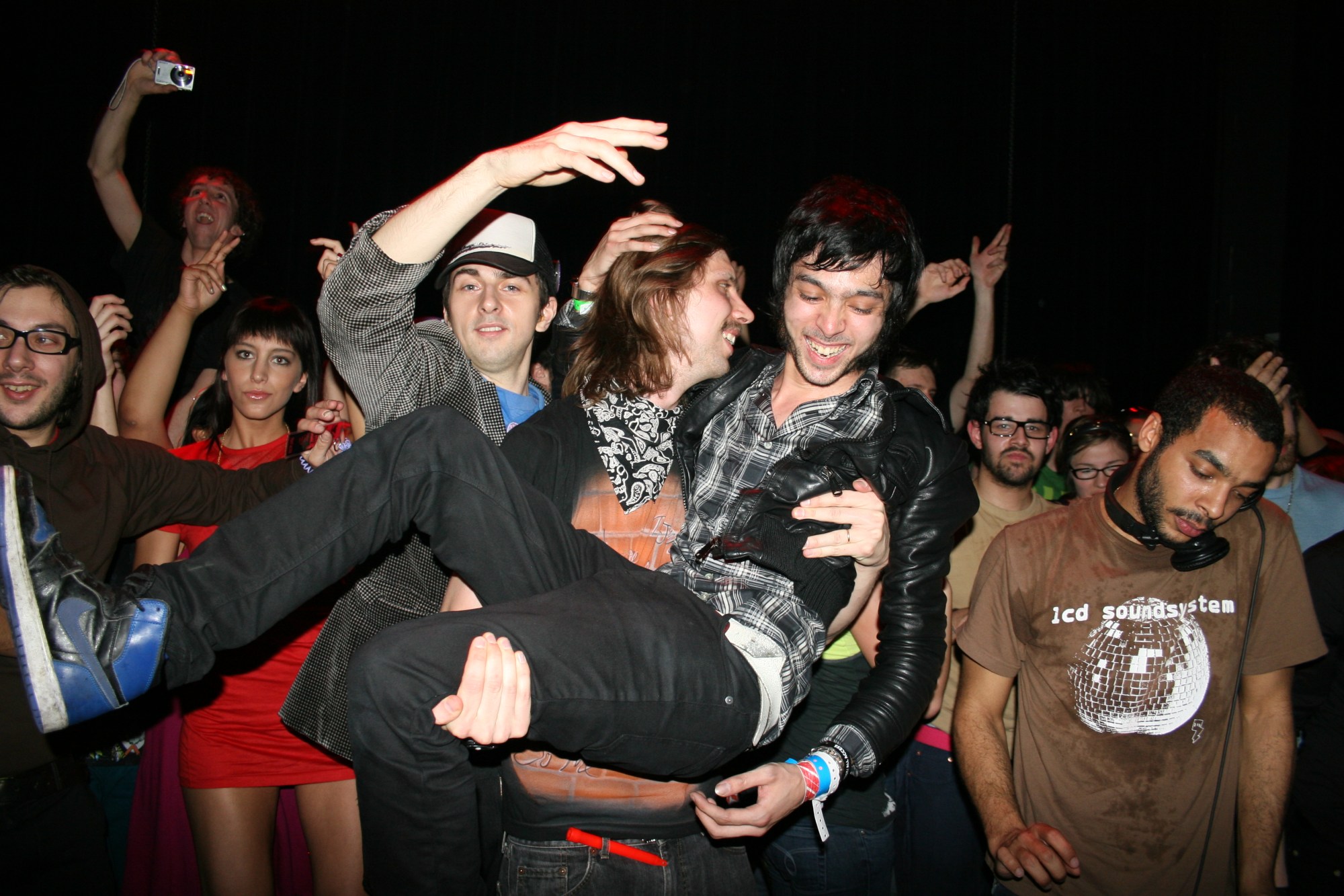 a man with long hair picking up his friend in front of a party of people photographed by the cobrasnake