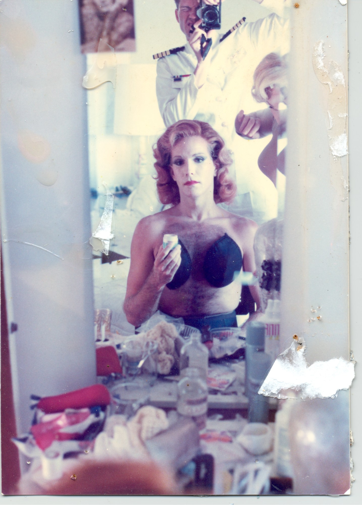 Person in a bra sitting at a mirror applying makeup as a sailor takes their picture through the reflection.