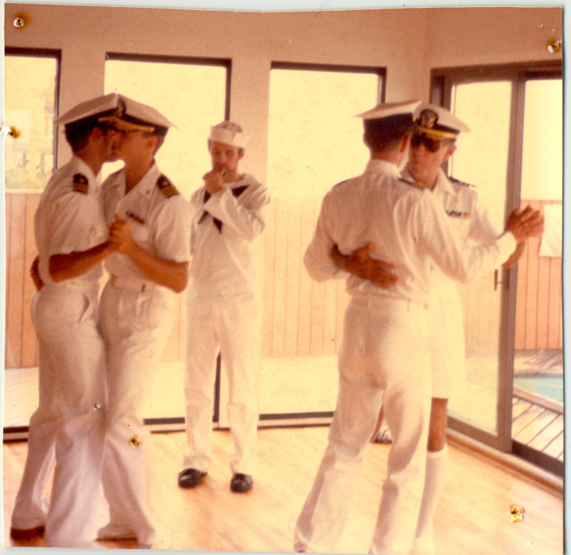 gay sailors dancing together in couples in a room on a boat deck whilst one stands on his own at the back.